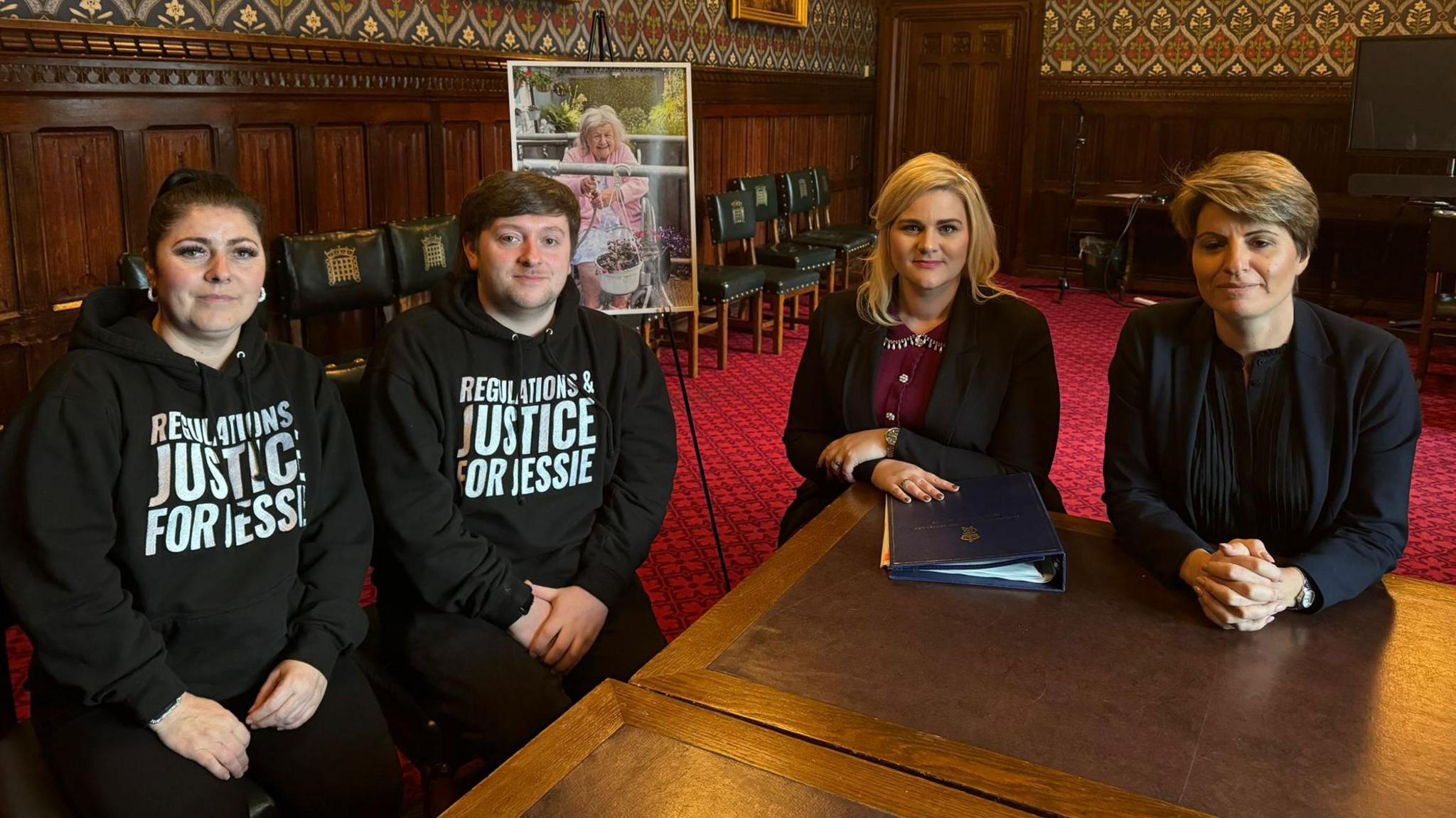 Four people sat around a table in a grand old room. Claire and Tristan are both wearing black hoodies with regulations justice for Jessie written on them. Next to Tristan is Alex Davies Jones, who has blonde hair and is wearing a black suit with purple top. Emma Hardy is sat wearing a dark blue suit and has her hands on the desk