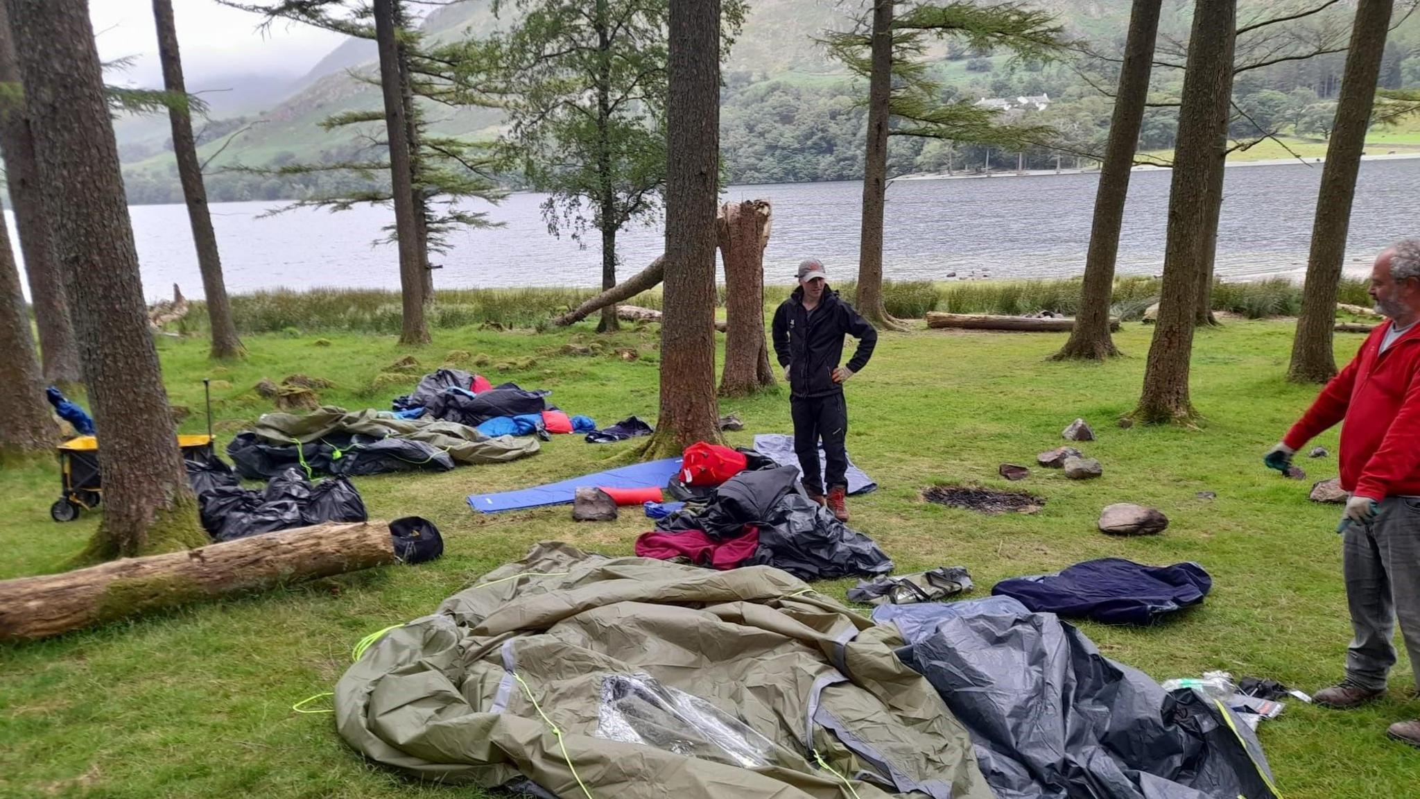 Two members of the National Trust stand at the lakeside at Buttermere with a discarded green tent in the foreground. Other items of camping equipment are strewn on the grass along with several black bin bags and items of clothing. There are several trees in the area and some have been chopped down.