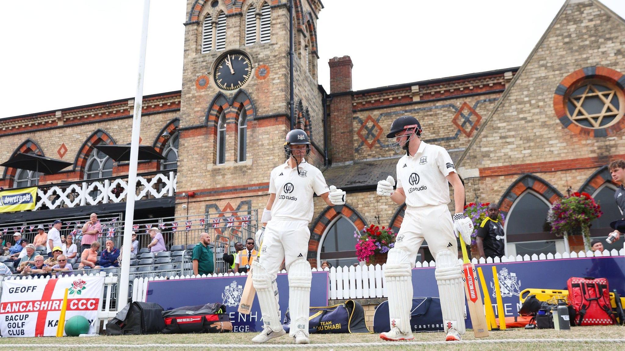 Two batsmen touch gloves as they walk over the boundary edge as Gloucestershire play Lancashire at the Cheltenham Cricket Festival In the background is the brickwork and large clock of the college building