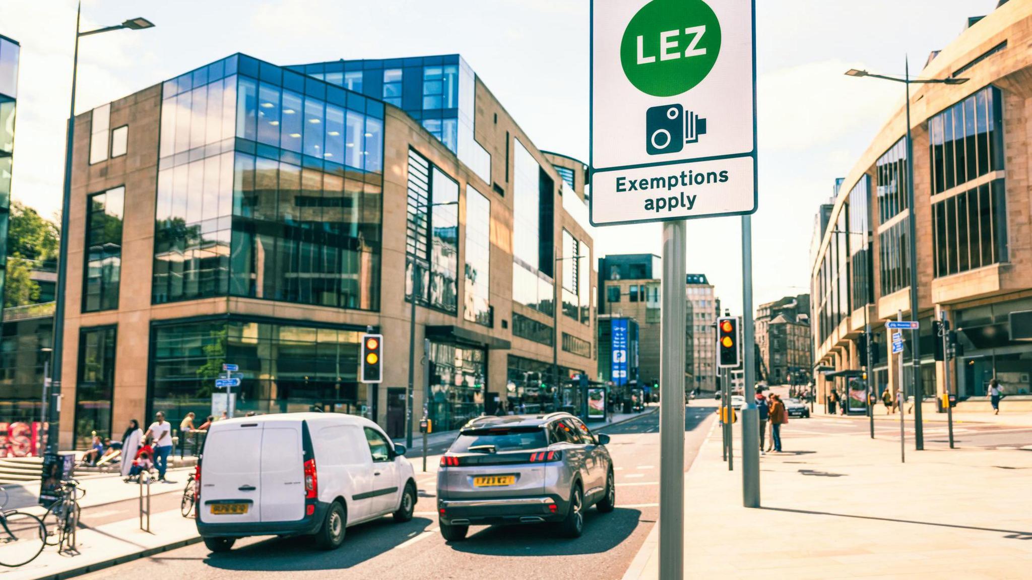 a sign that reads low emission zone exemptions apply on a road in edinburgh with two cars waiting at traffic lights nearby and pedestrians walking
