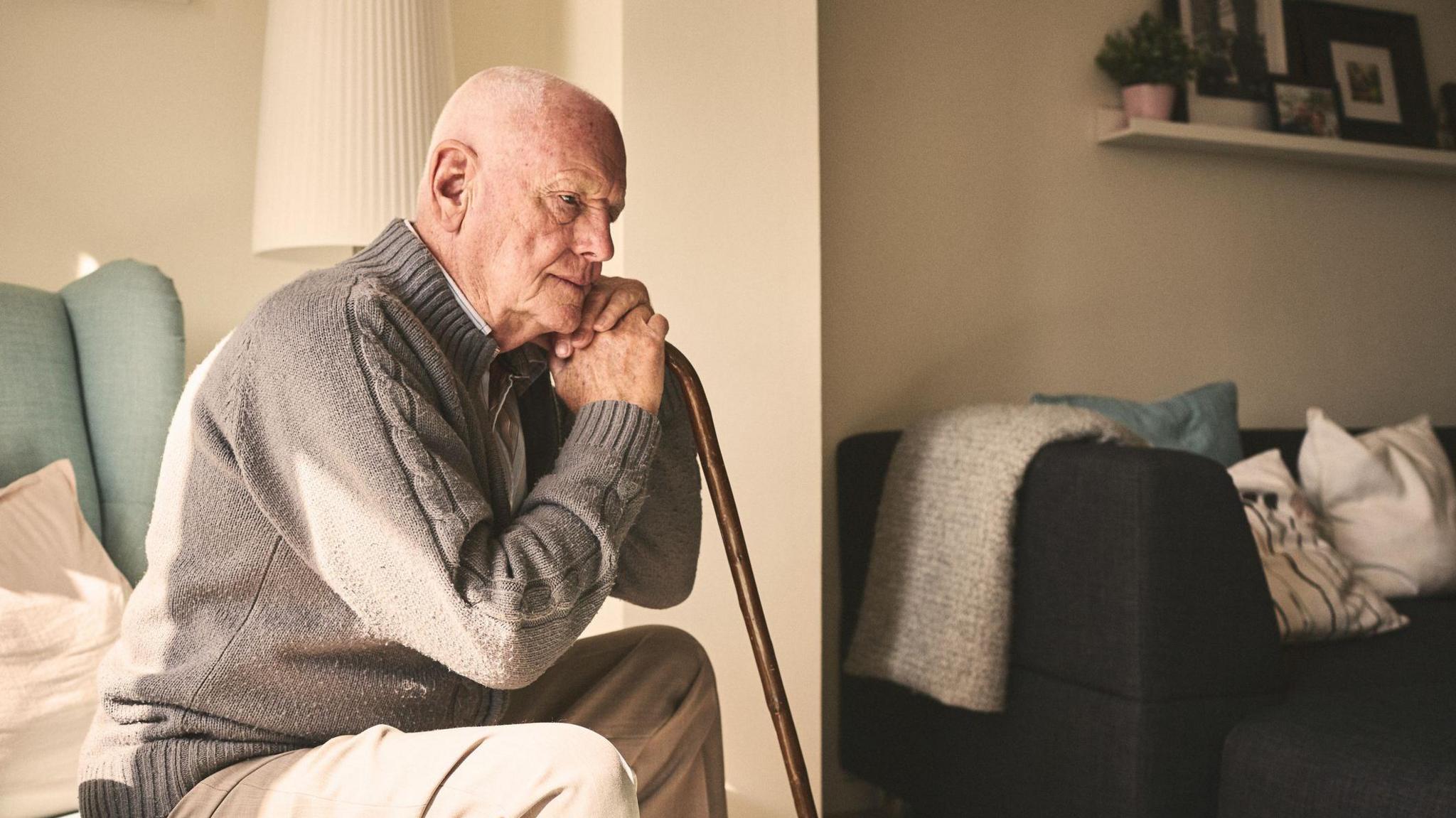 An elderly man in a brown zipped cardigan sits on a green chair in a care home, leaning against his stick. There is a lamp and another sofa with cushions in the background as well as a shelf containing plants and pictures.