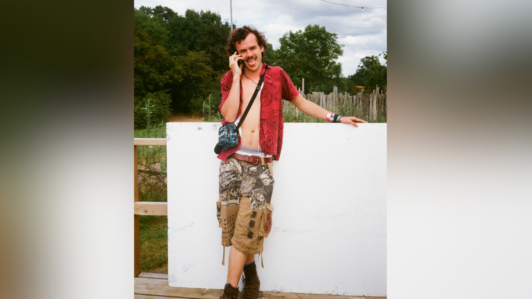 Woody Cook stands in front of a white wall. He's wearing an open red shirt and camouflage shorts