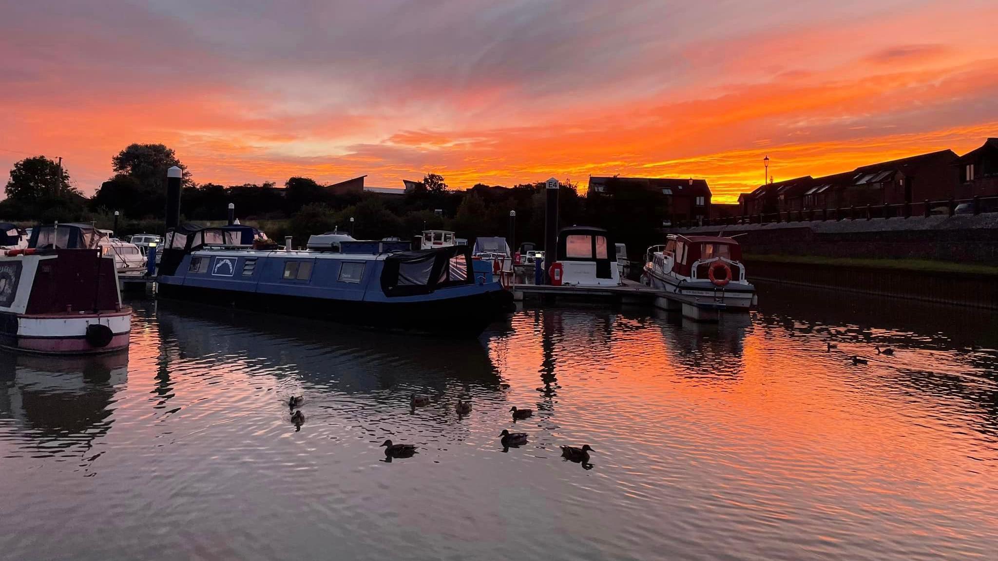 A sunrise is pictured over houseboats on water in Tewkesbury. The sky is a mixture of red and orange and is reflected in the water, which also has ducks sitting on it