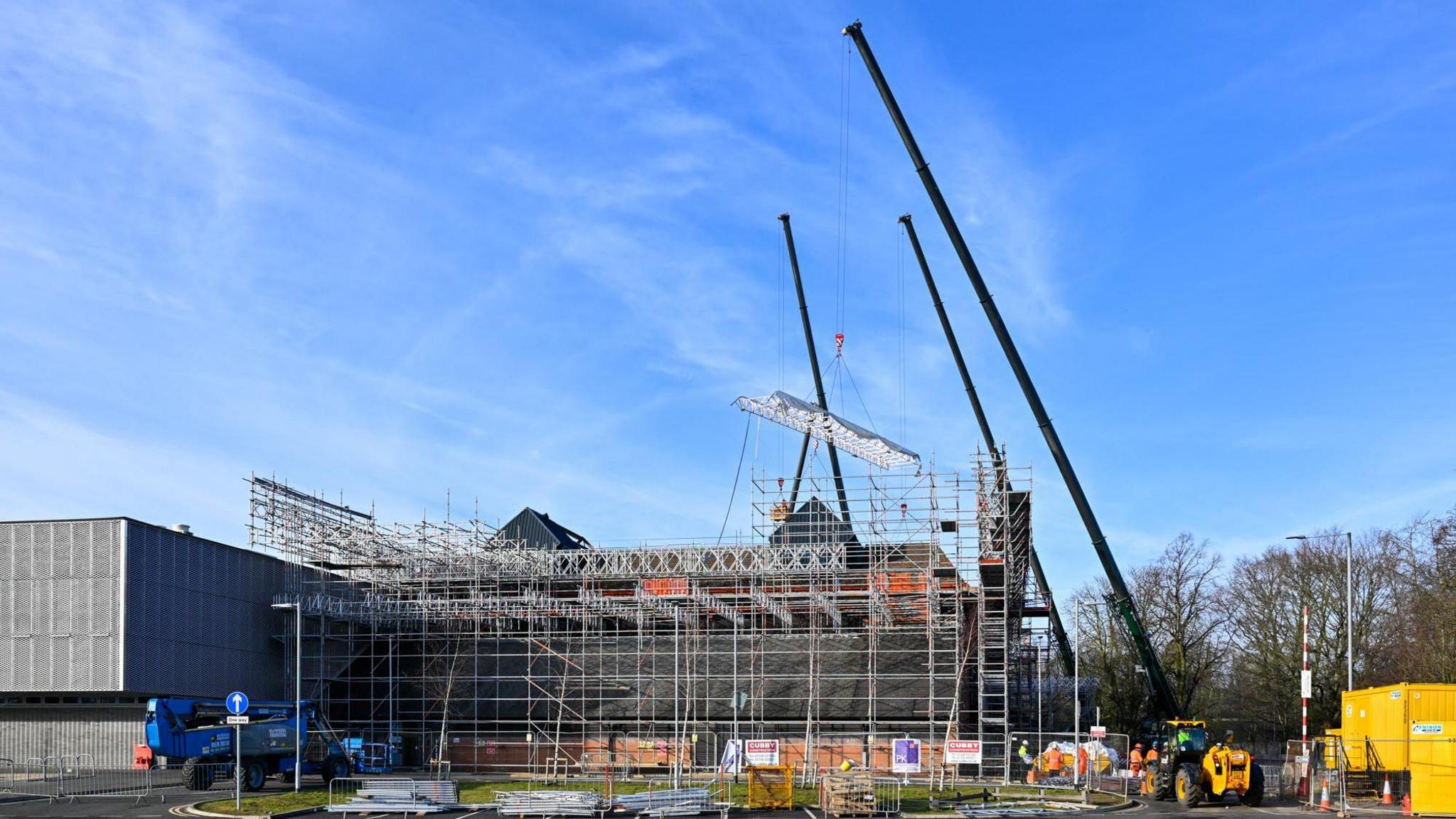 The Sands Centre which is a grey building covered with scaffolding. Three black cranes are beside it.