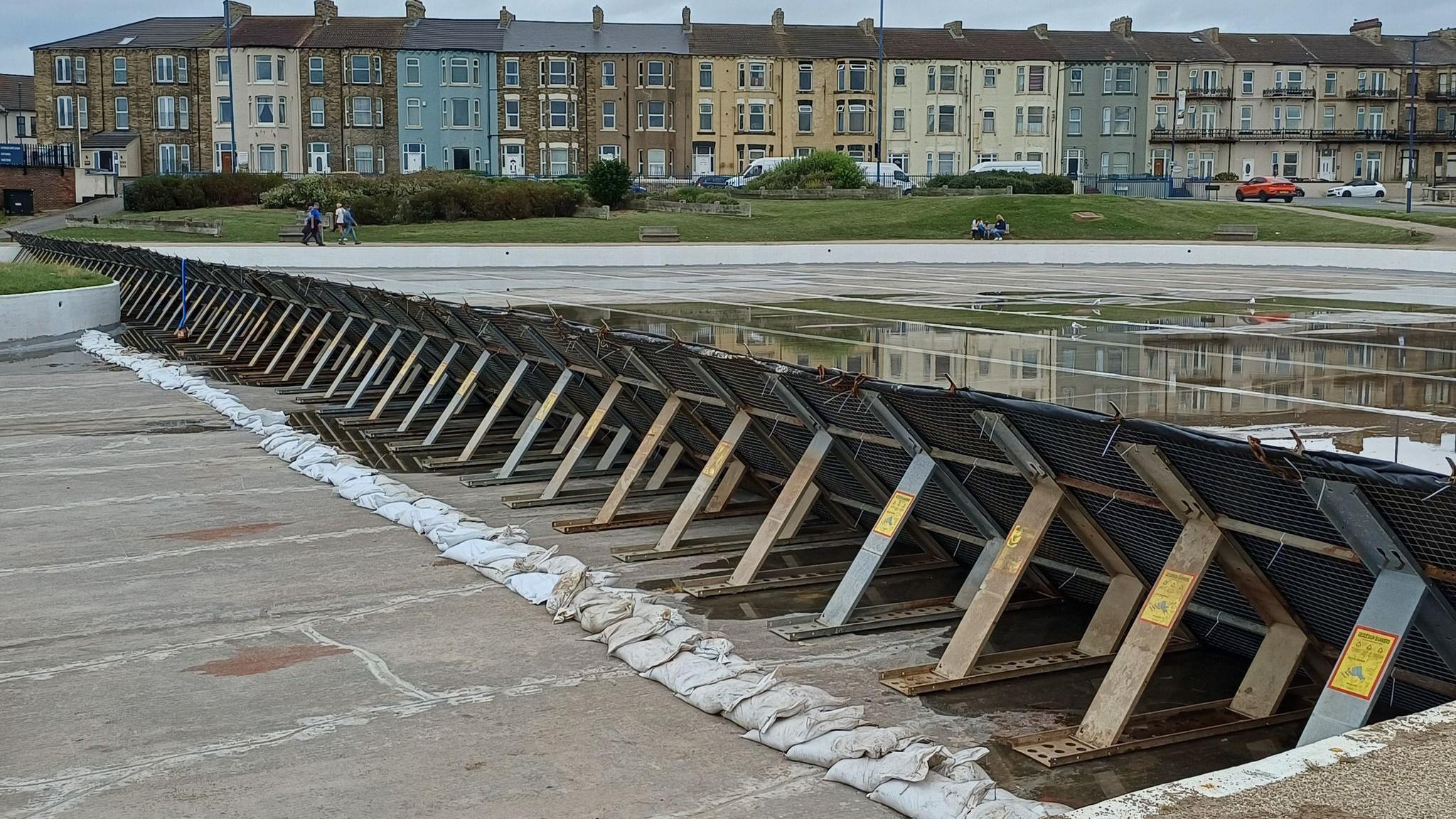 A dam with sand bags is stretching along the middle of the lake. There is a row of three-storey houses across the road. Several people are walking past. 