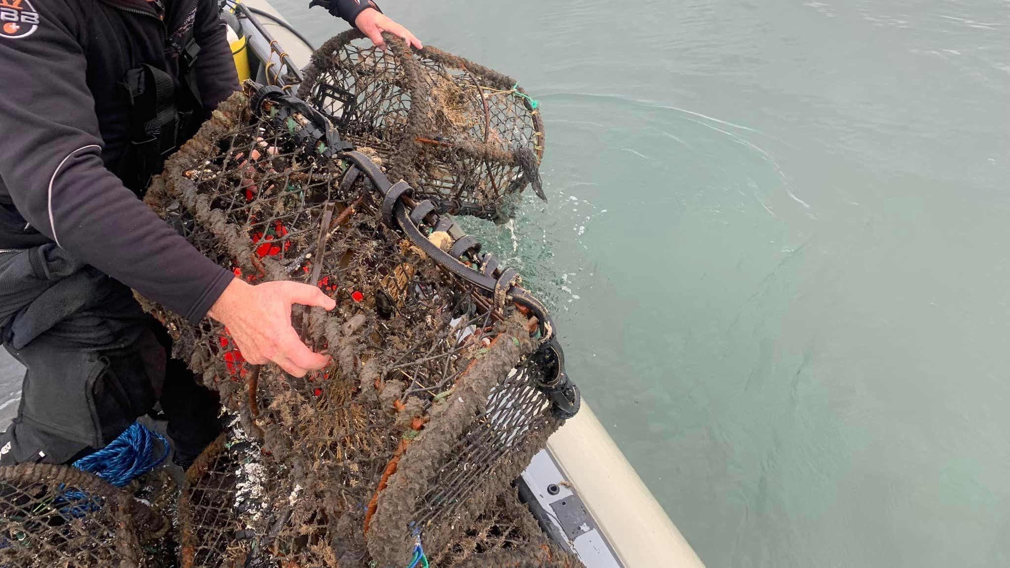 Two hands holding a lobster pot over the edge of the sea on a boat. The pot is large and brown. 