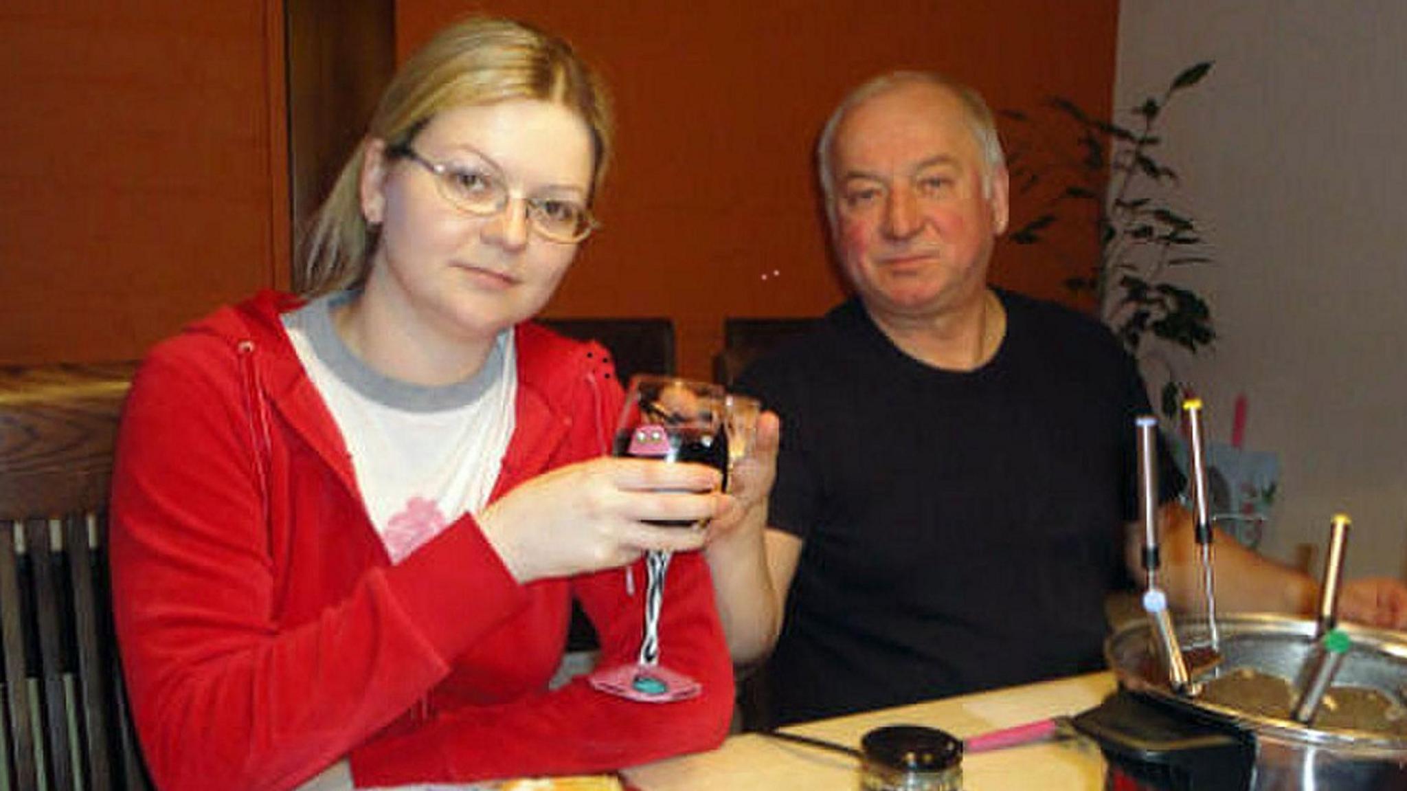 Yulia and Sergei Skripal. Yulia has blonde hair which is tied in a ponytail. She is wearing glasses, a white t-shirt and a red zip hoodie. She is sitting next to her father Sergei, who has short grey hair and is wearing a black t-shirt. They are both sat at a table, holding glasses full of drink. They are looking at the camera and smiling.