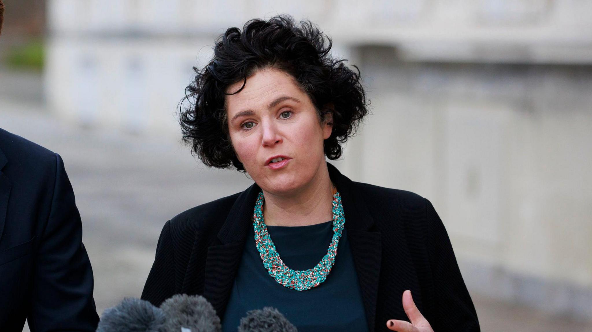 Claire Hanna standing speaking at microphones outside parliament buildings at Stormont. She is wearing a blue top, navy jacket and blue necklace.