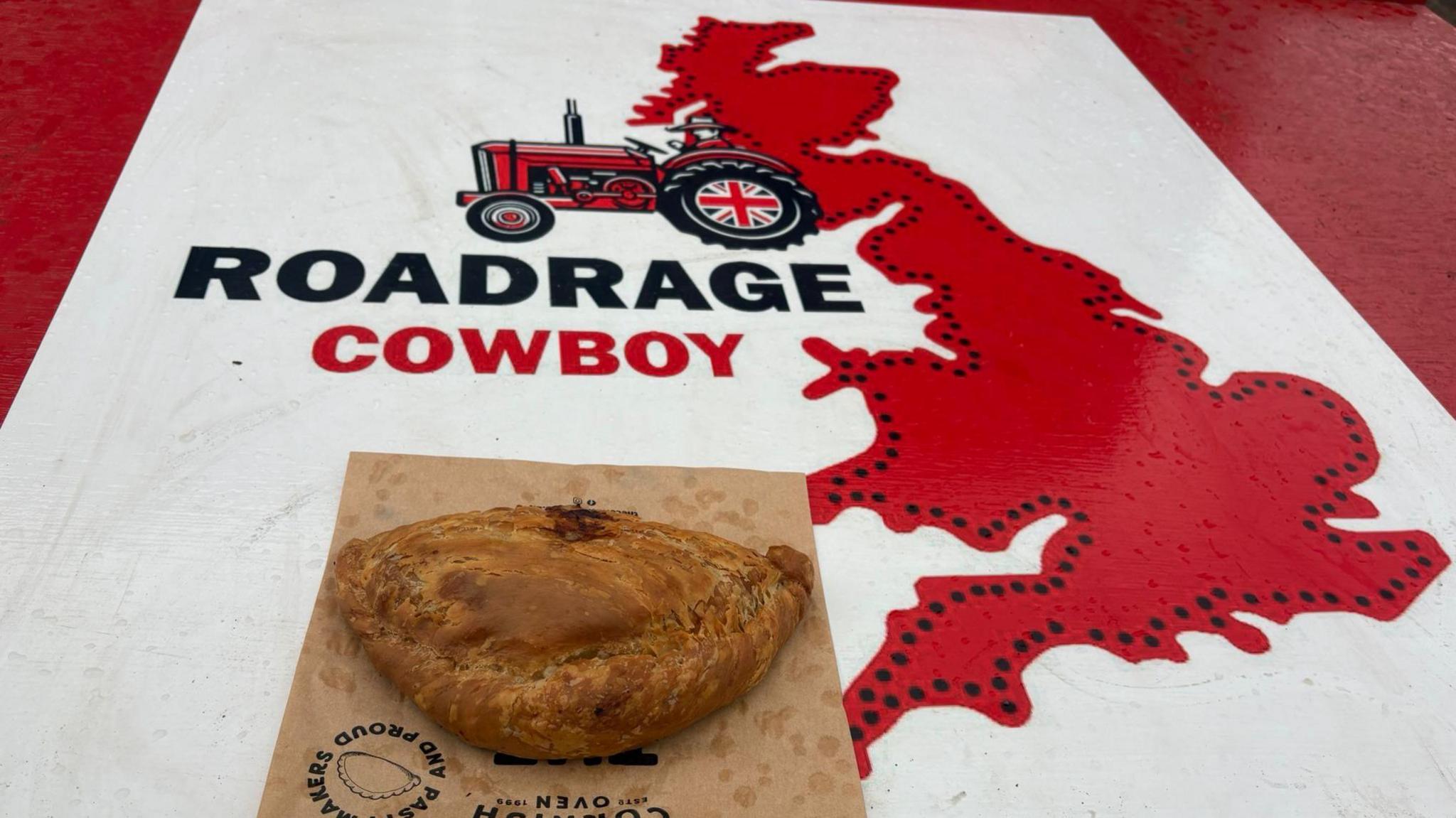 A Cornish Pasty on top of the hood of a tractor. The hood has "Roadrage Cowboy" written on it with a picture of a red tractor and Britain.