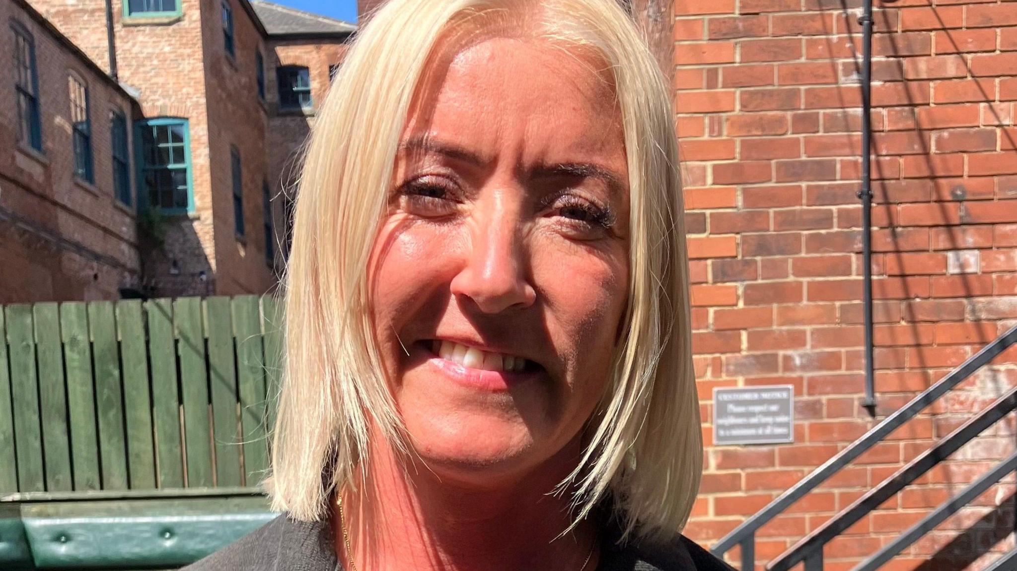 A blonde woman smiles at the camera. She is standing in a pub garden