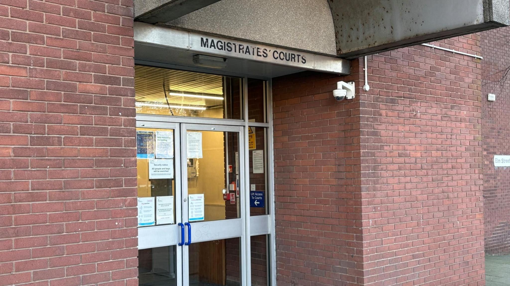 The front of a red brick building with double glass doors is seen, with the words 'Magistrates' Courts' pictured above the glass doors