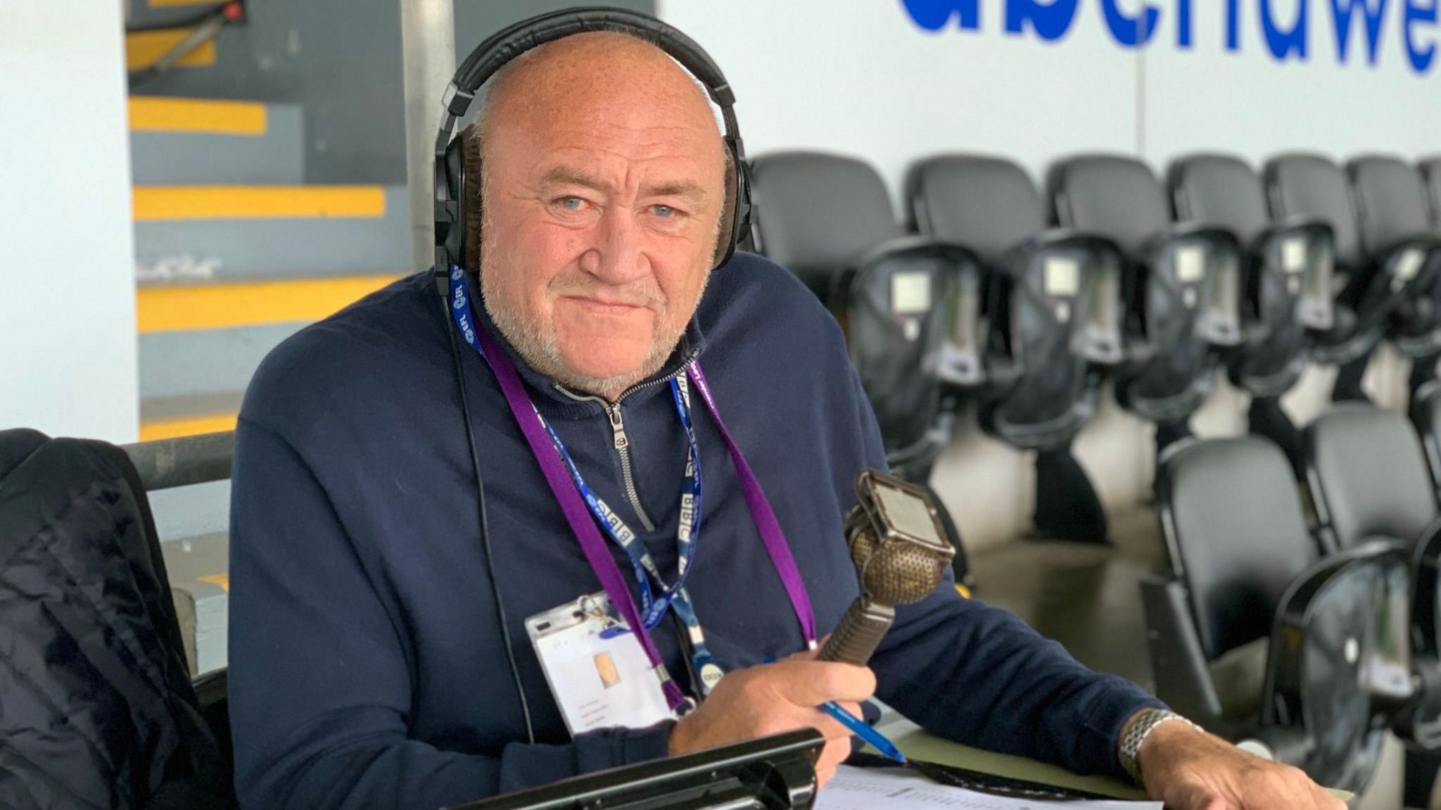 A man looks into the camera with a serious expression. He has is wearing headphones and around his neck is a navy blue lanyward with the BBC logo on it in white. In his right hand he is holding a radio microphone. Behind him are rows of stadium chairs in the upright position.