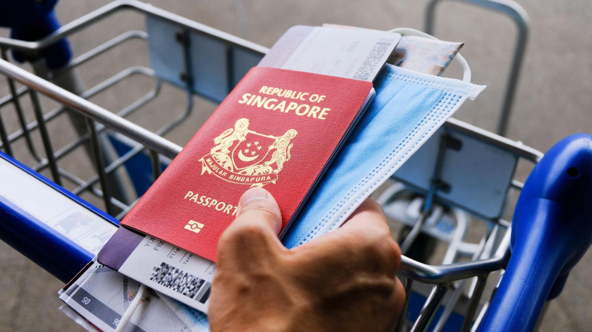 In airport: hand on luggage trolley, holding Singapore passport, boarding pass, face mask, and map. 