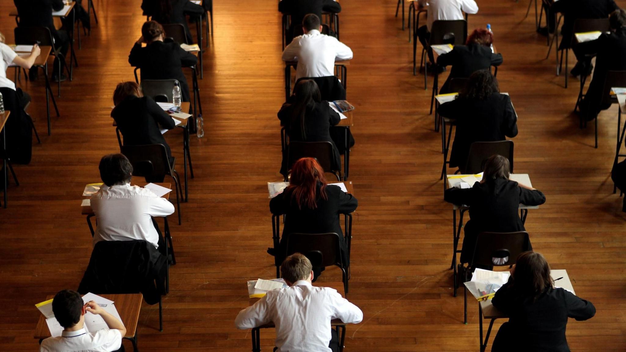 View from behind of pupils doing exams in a school hall. They are sitting at separate desks which have been spaced out for exam conditions. There are papers on their desks. Some of them have got black jumpers on while others are just in white shirts.