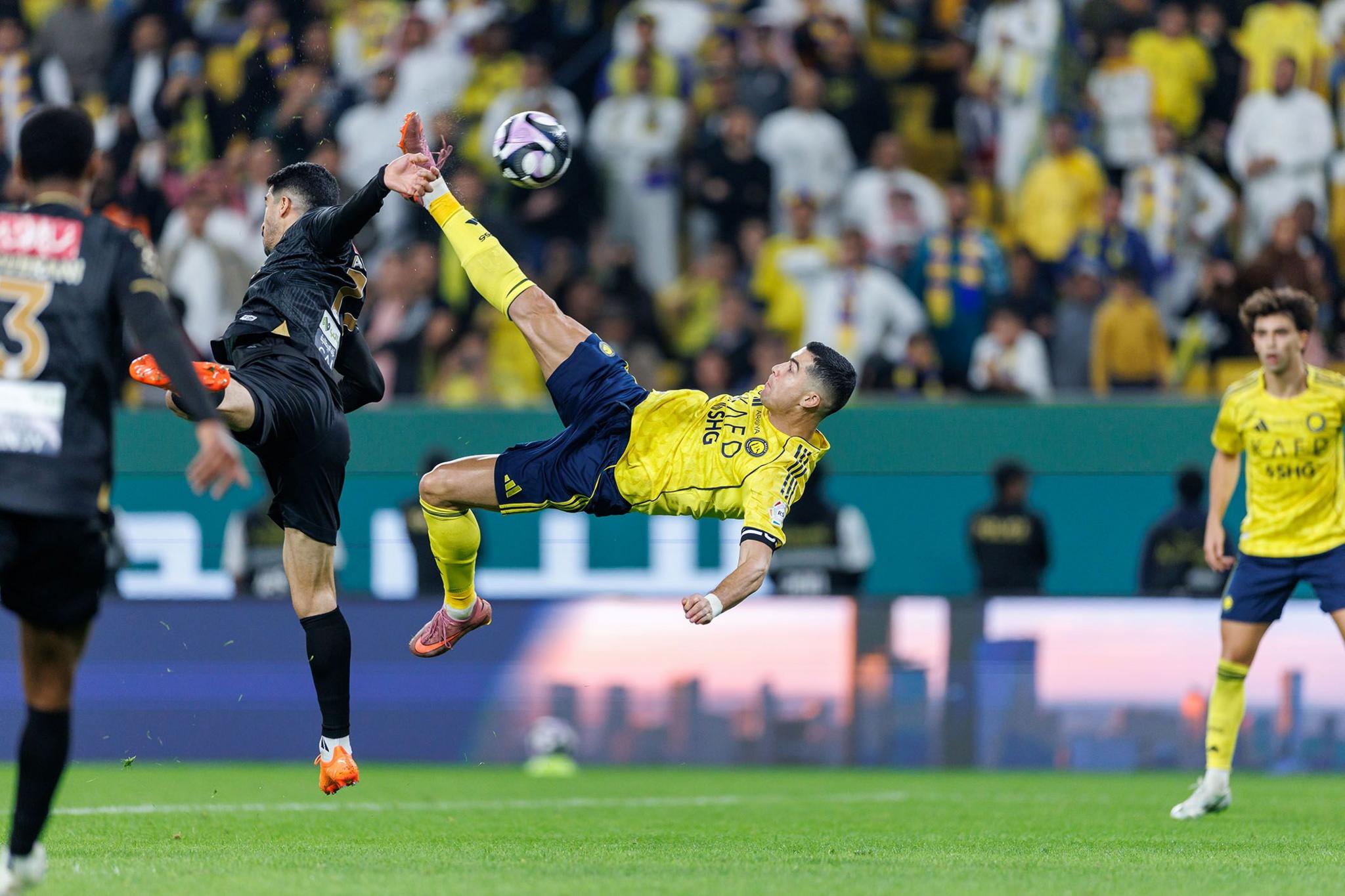 Football player in a yellow jersey performing an acrobatic bicycle kick during a match, with an opponent in black attempting to block and a crowd of spectators in the background.