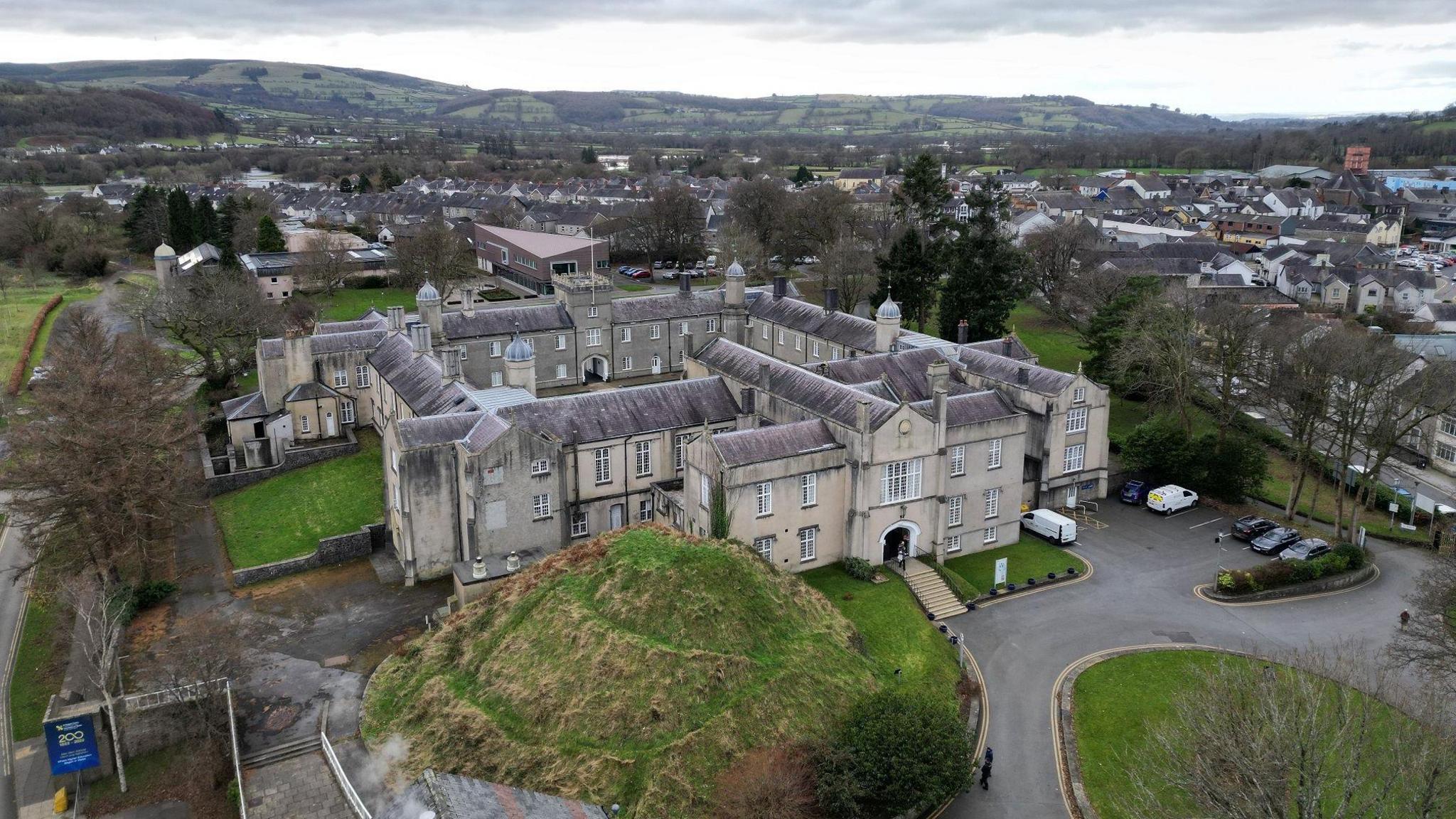 A drone picture of the university. It shows the entrance of the historic stone building next to a large grass mound, with the inner courtyard behind. The town of Lampeter can be seen on the right hand side.