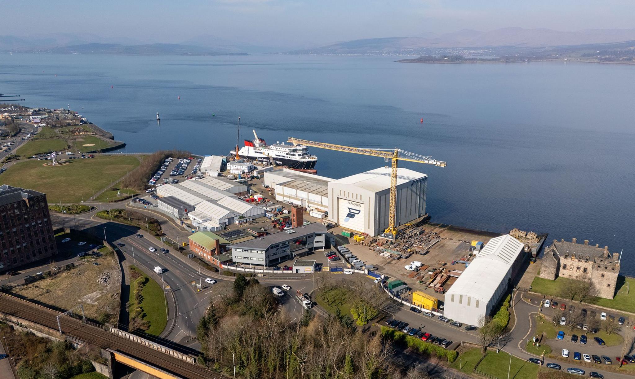 An aerial view of a shipyard. There are grey buildings, a yellow crane and a large ship berthed on the quayside