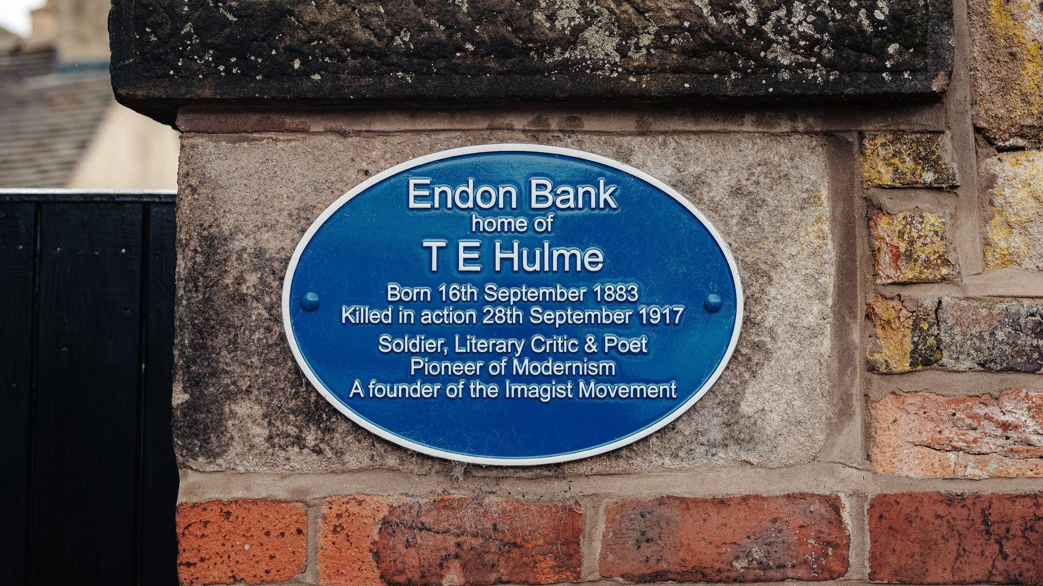 A blue plaque on a brick wall is dedicated to TE Hulme. It is oval shaped with white writing, detailing the dates of his birth and death as well as his credentials as a poet.