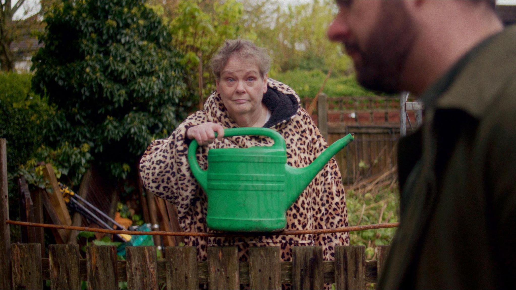 A shot of Anne Hegerty from the film. She is holding a green watering can and wearing a leopard print hooded top. She is standing in a back garden. She is looking mischievously at a man slightly out of shot.