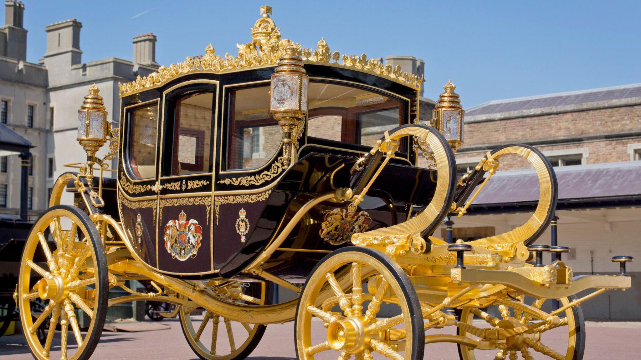 A modern coach with gold wheels and a painted main structure. It sits in front of the Royal Mews. 