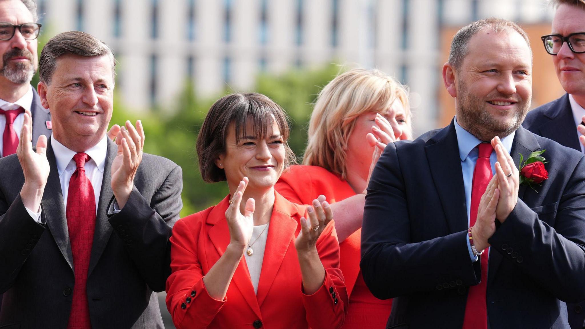 Alexander and murray in suits clap their hands with a woman in a red suit between them.