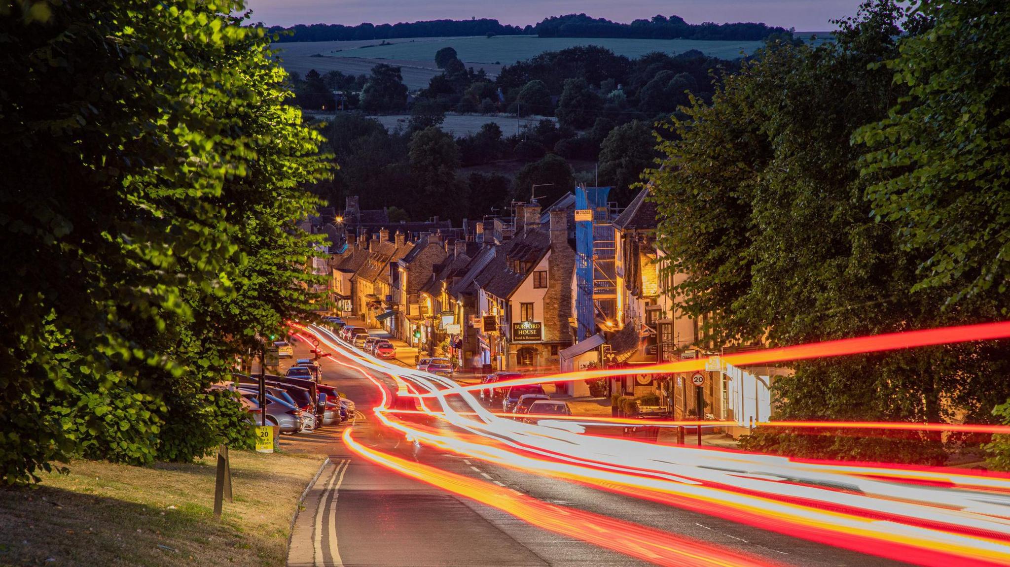 Burford is a town in the Cotswold hills, Oxfordshire, United Kingdom. This photo shows high street traffic at night in Summer 2022