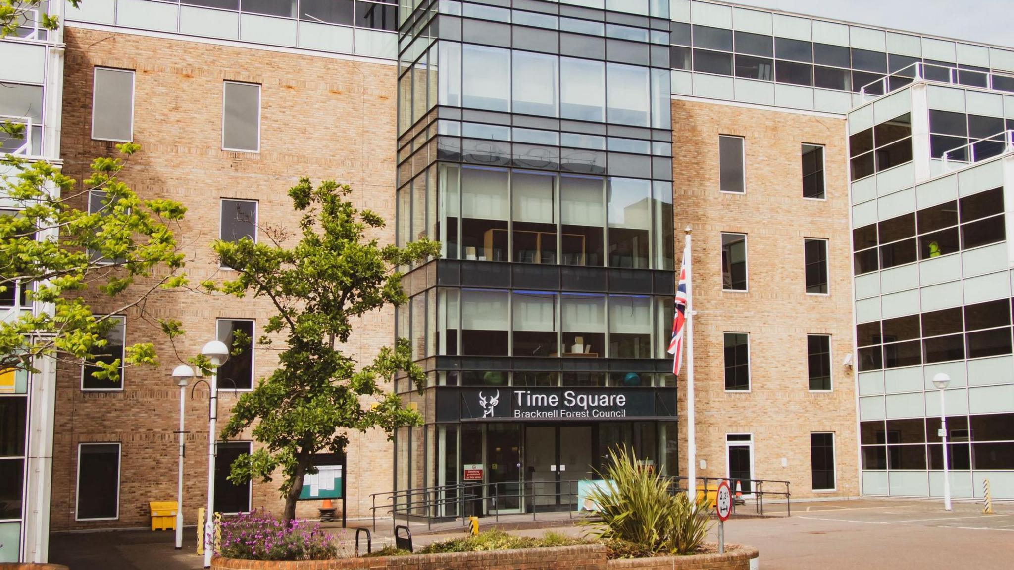 The Time Square Offices of Bracknell Forest Council in Market Street, Bracknell, taken in May 2020. It has five floors, with four of them on the left and right side having orange bricks. But in the middle of the building, there is a glass-fronted centre, which has five panes of large glass across and is five storeys high. It has "Time Square, Bracknell Forest Council" written on a sign above the entrance, next to the the council's logo of a deer