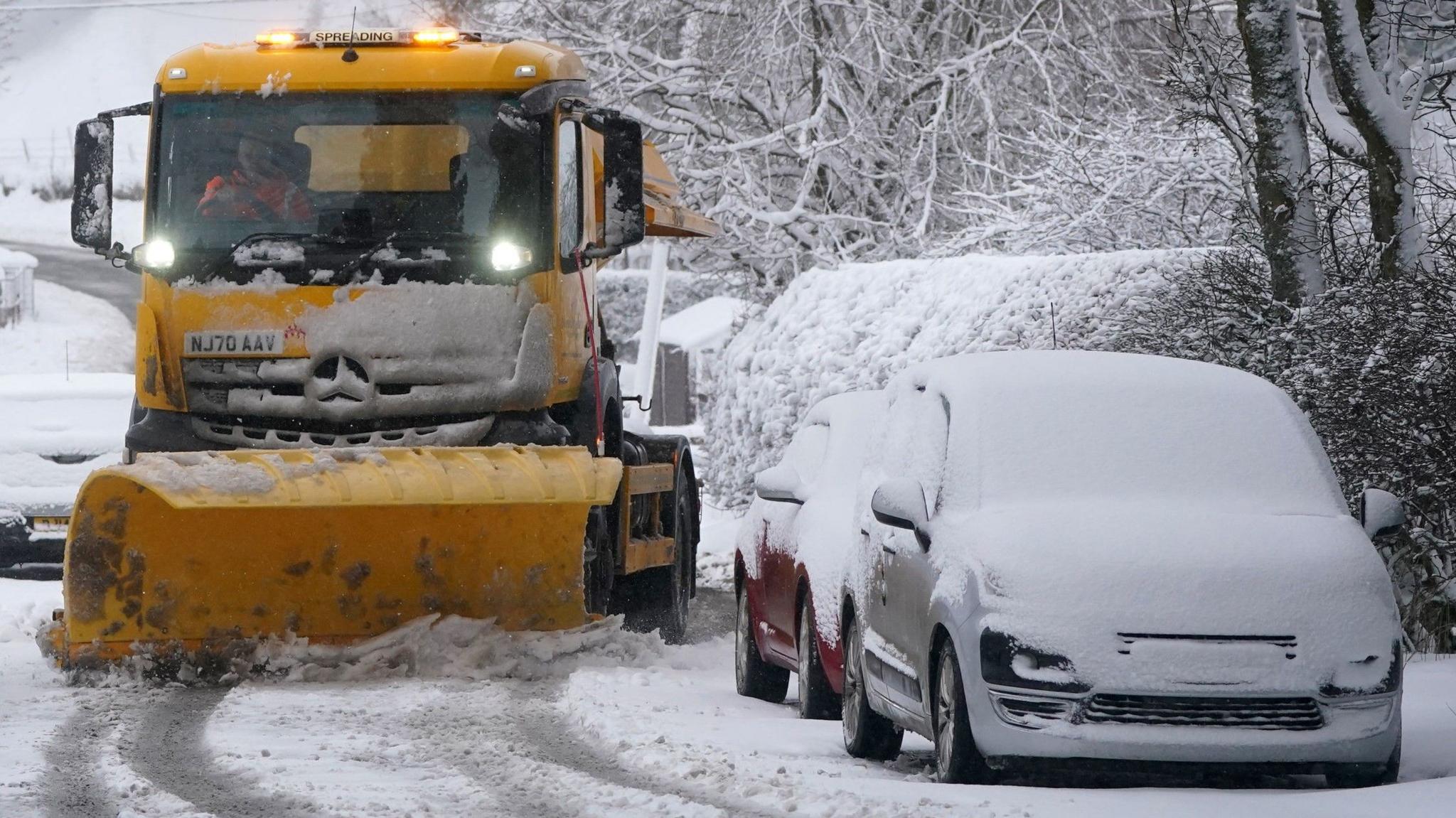 A snow plough manoeuvres past two snow-covered parked cars