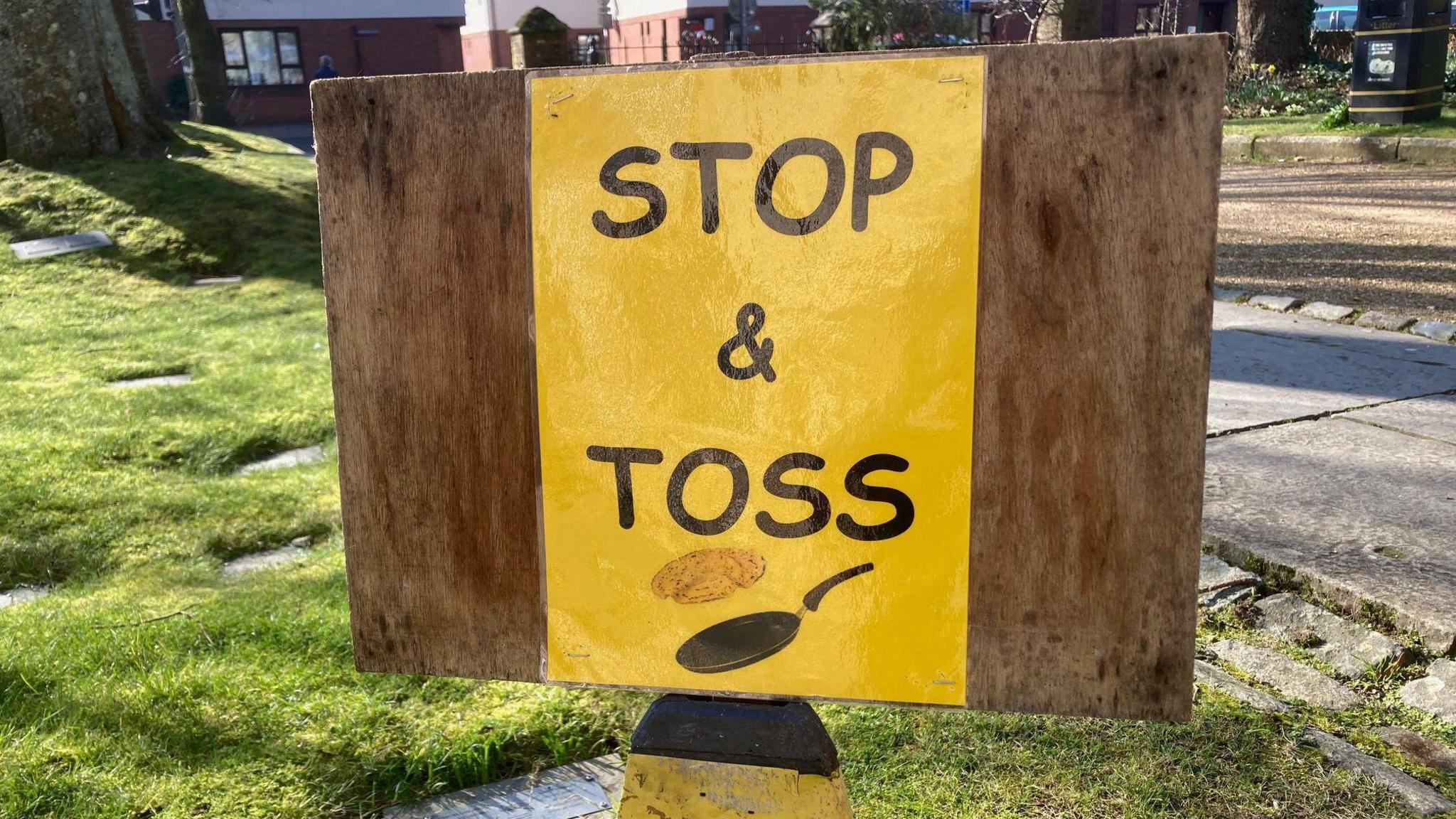 A wooden board sign, with a yellow piece of paper stuck to it reads, "Stop & Toss" on the race route