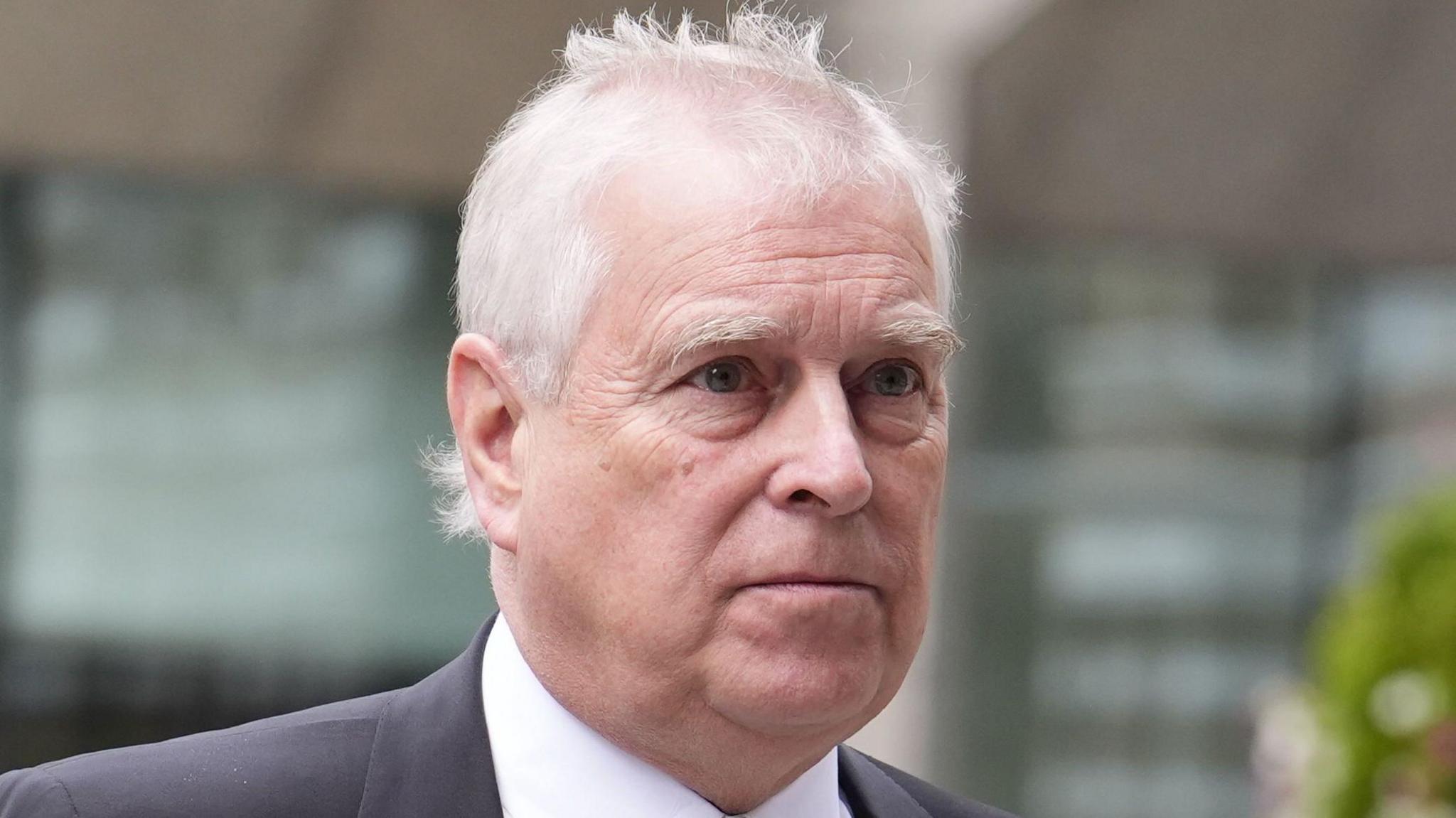 Andrew Mountbatten-Windsor, with short white hair in a black suit and tie, walks past a glass building
