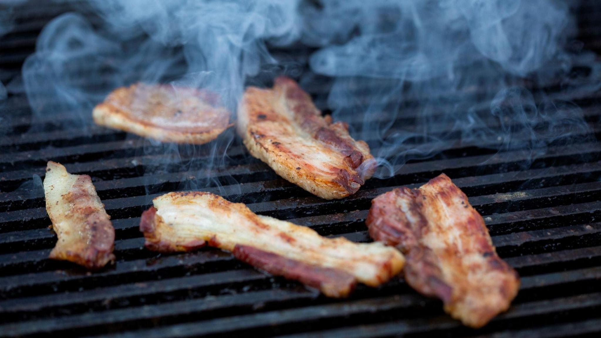 A stock image of five bacon rashers on a grill smoking