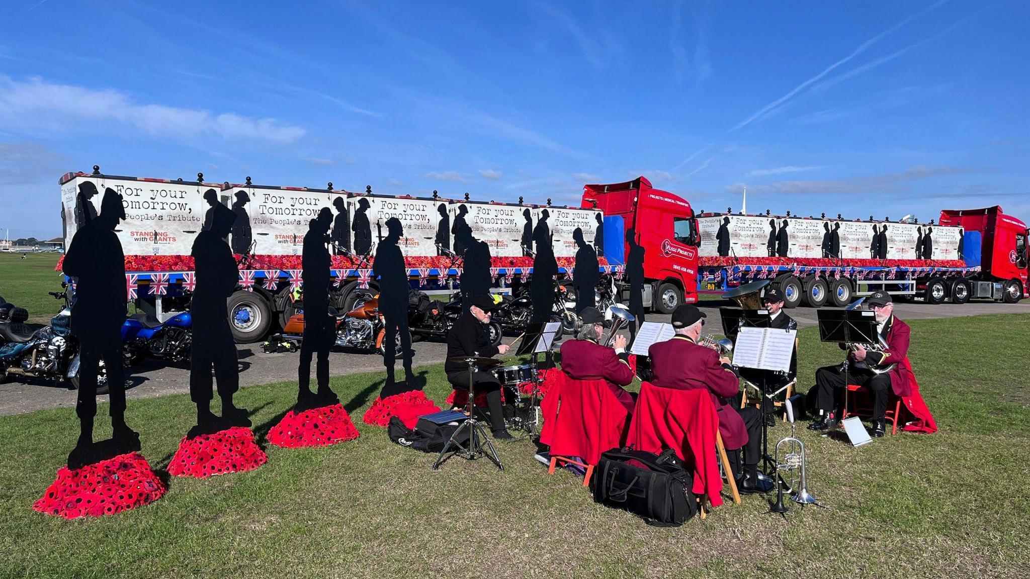 The silhouette statues with members of the military band playing music in front of them and large lorries behind them 