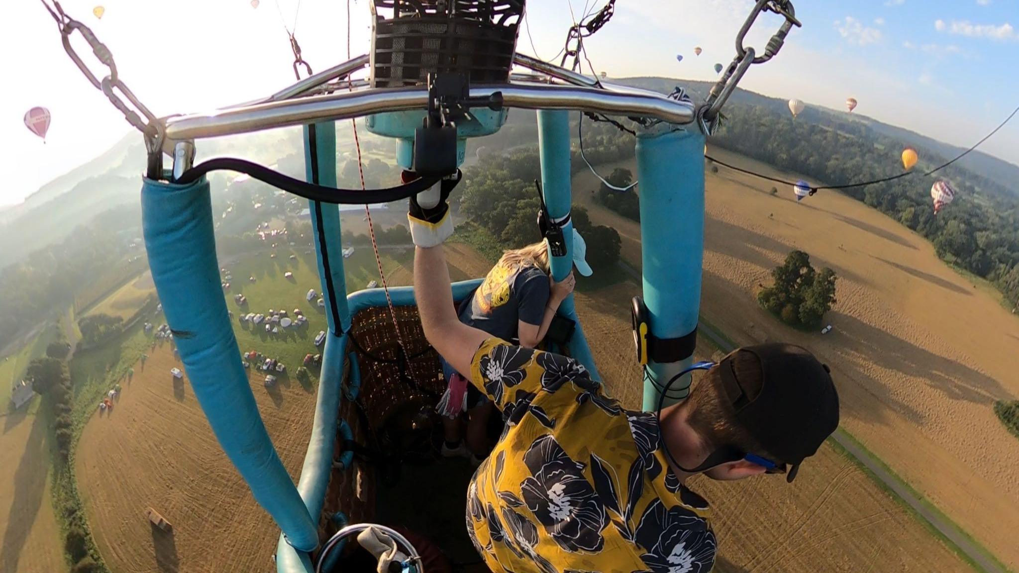 Max in a hot air balloon basket looking down on fields and other balloons below