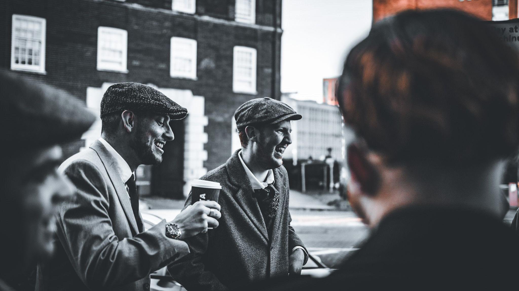 A group of men wearing flat caps and tweed suits laugh, whilst stood outside of a building. One man is drinking a coffee.
