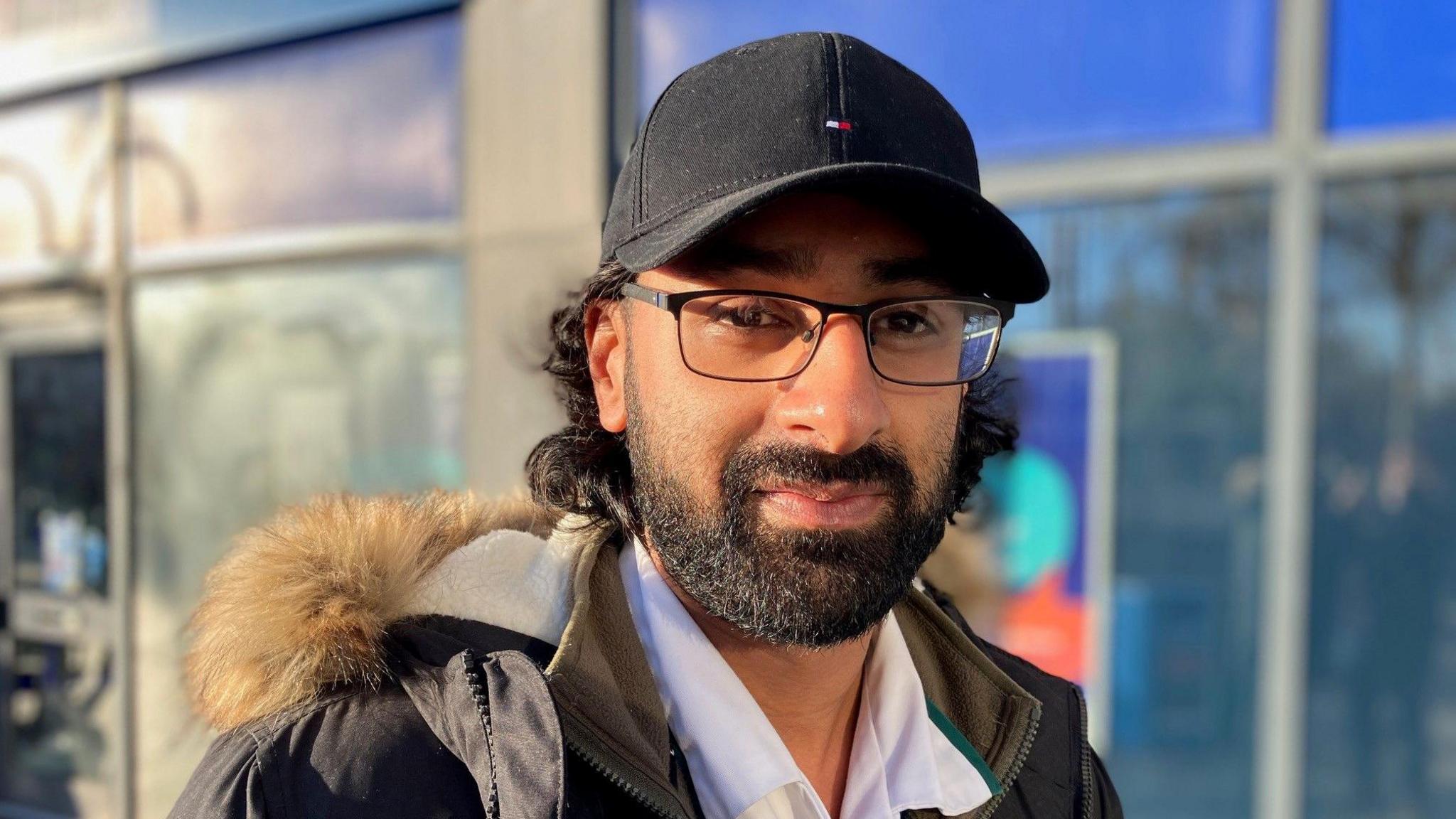 A man wearing glasses smiles while standing in front of shop windows. He has a beard and curly dark brown hair and is wearing a shirt, olive-coloured coat and black baseball cap.