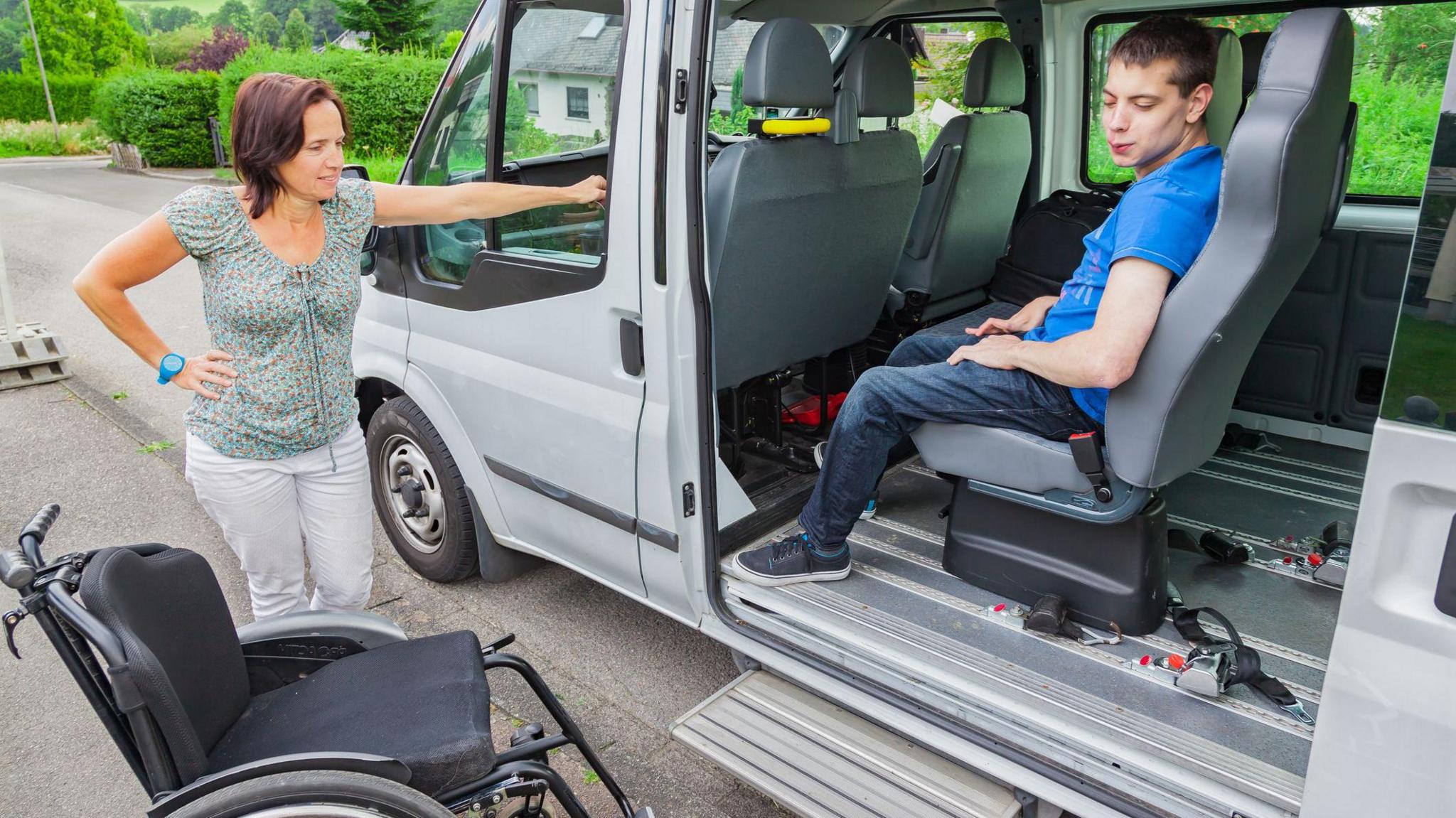 A boy in a blue t shirt sitting in a special needs school bus. A woman is standing to the left of the bus next to to a wheelchair