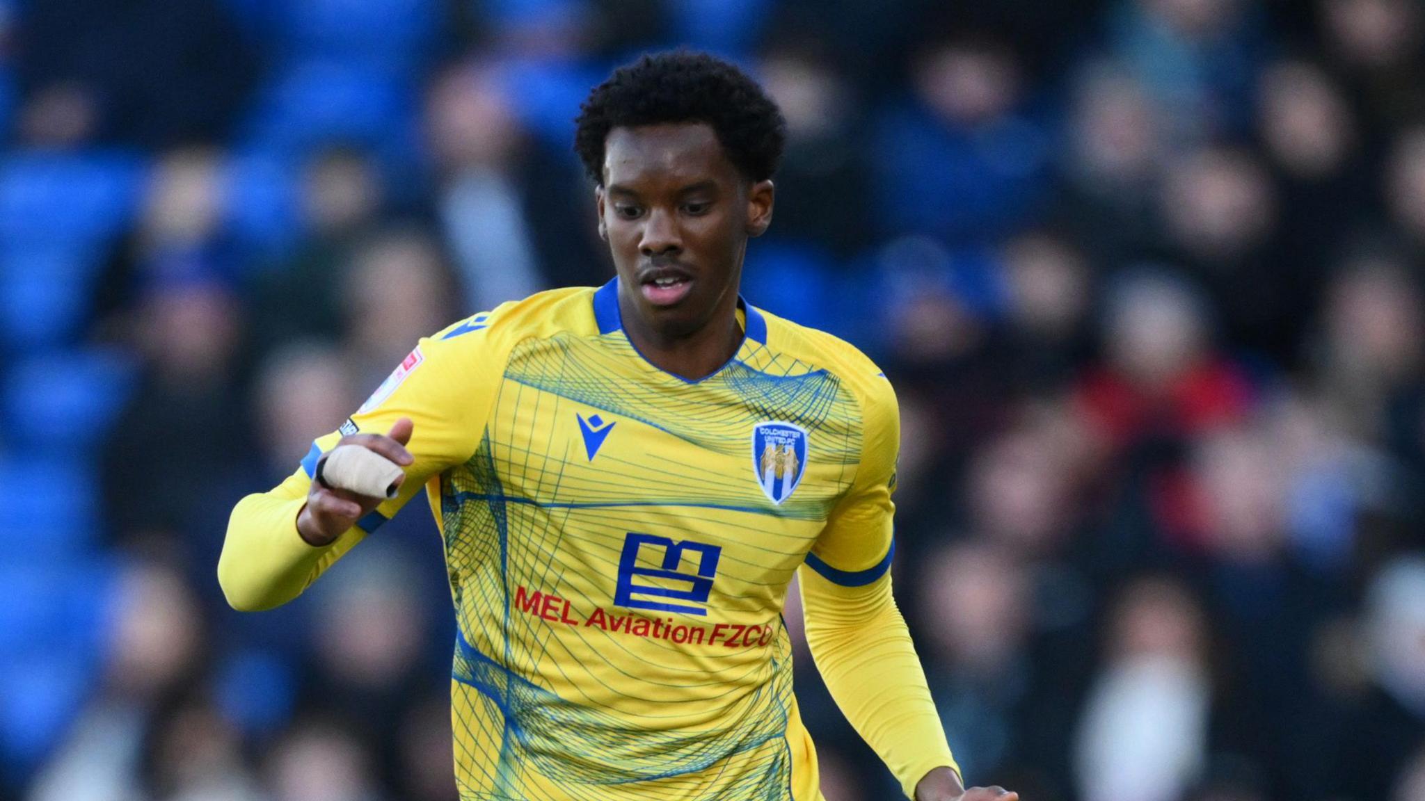 Harvey Araujo playing for Colchester United in their yellow kit, with two of his fingers taped together