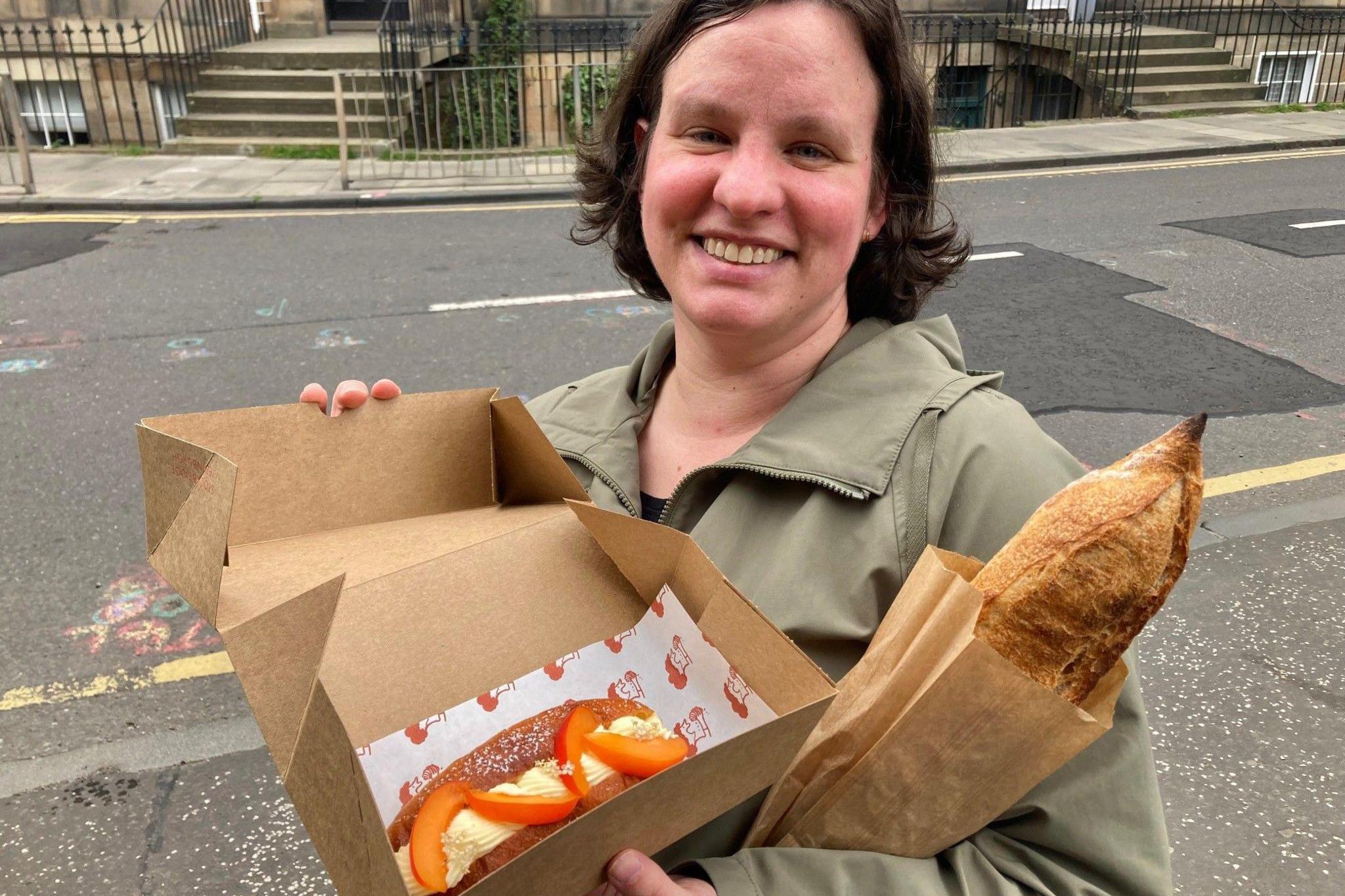 Sophia is smiling and holding a cake box open with a cake inside. she has brown hair and is wearing a green coat. She has a stick of bread under her arm.