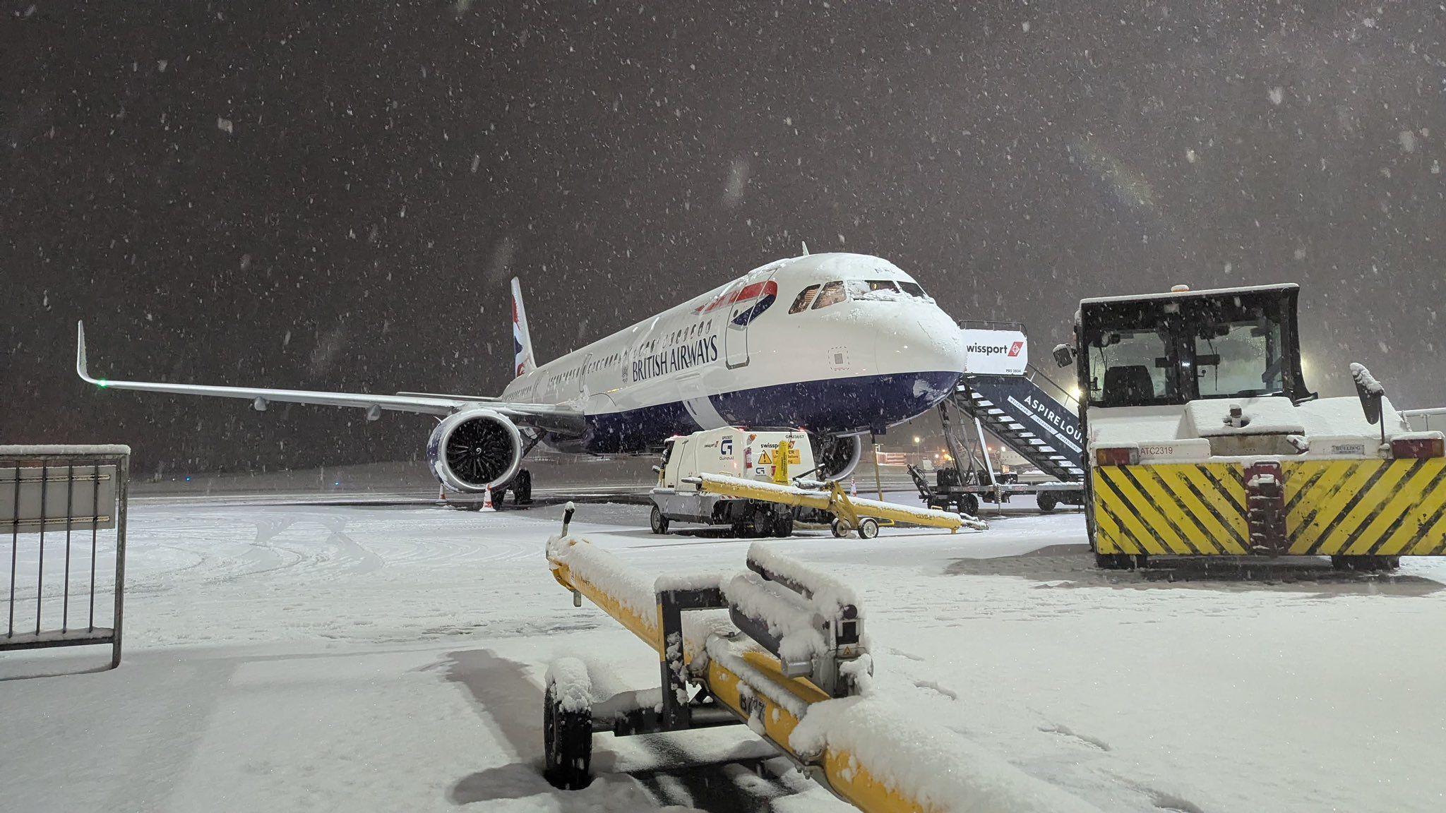 A British Airways plane is on the runway as snow falls around it. Snow blankets the ground.