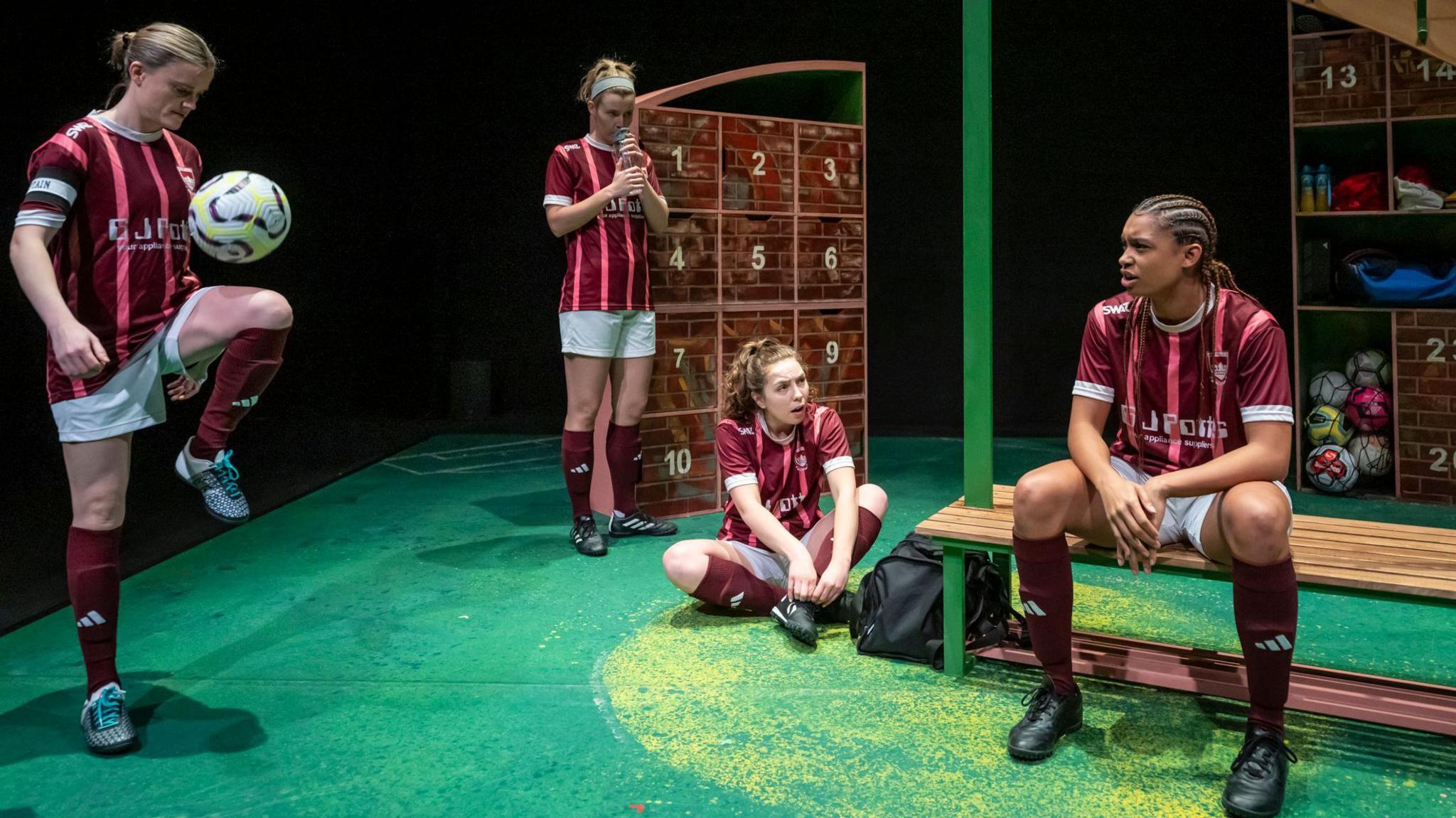 Four women in a red and white football kits are in a stage set up of a changing room. The woman on the far left is kicking a football up with her knee, another is standing by a locker, a third is sat cross-legged on the fourth and the woman on the right is sat on a bench