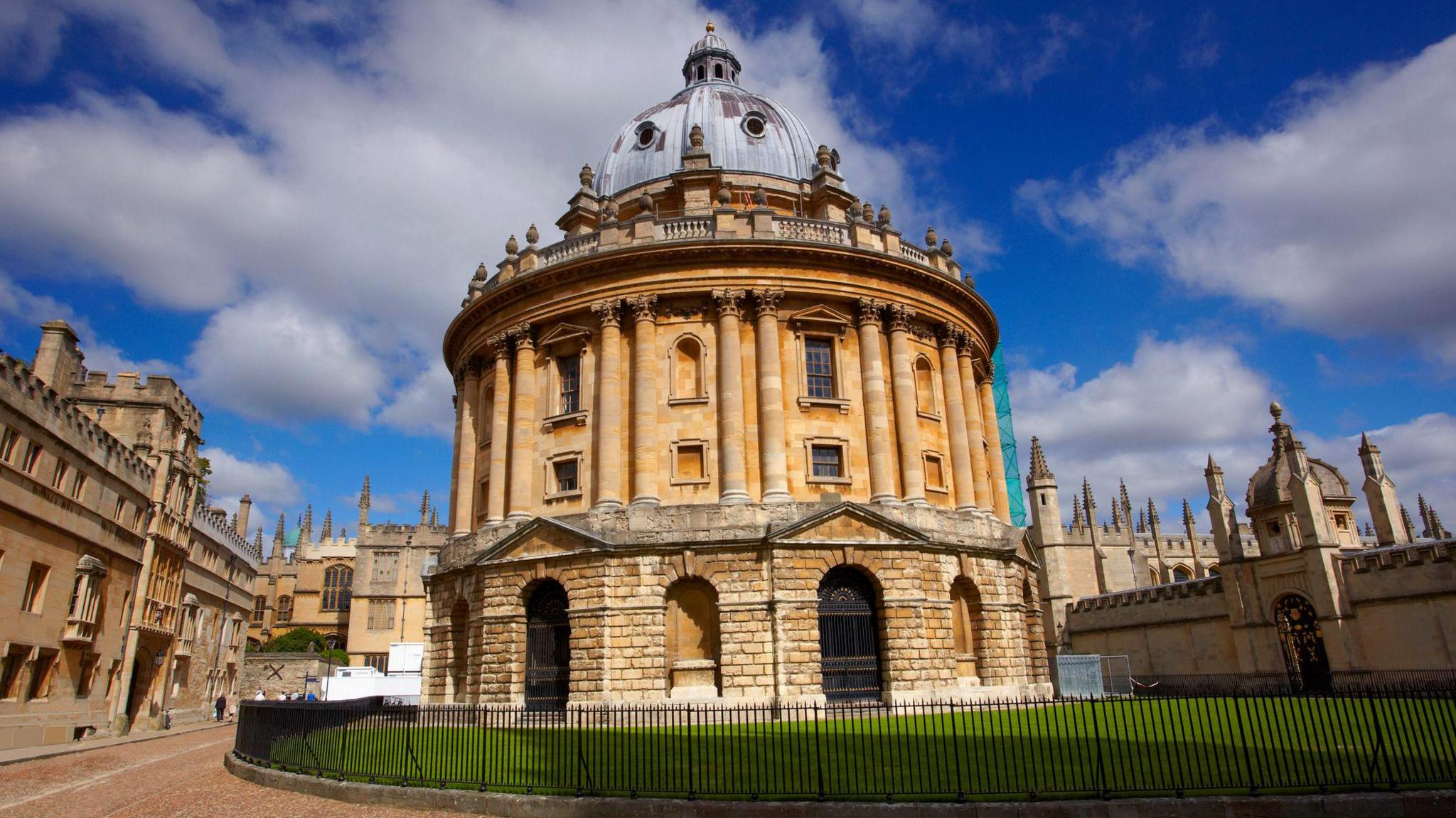The Radcliffe Camera, a circular building with pillars and a domed roof. There's grass next to it and a black railing running round it. It's a sunny day and the sky is blue.