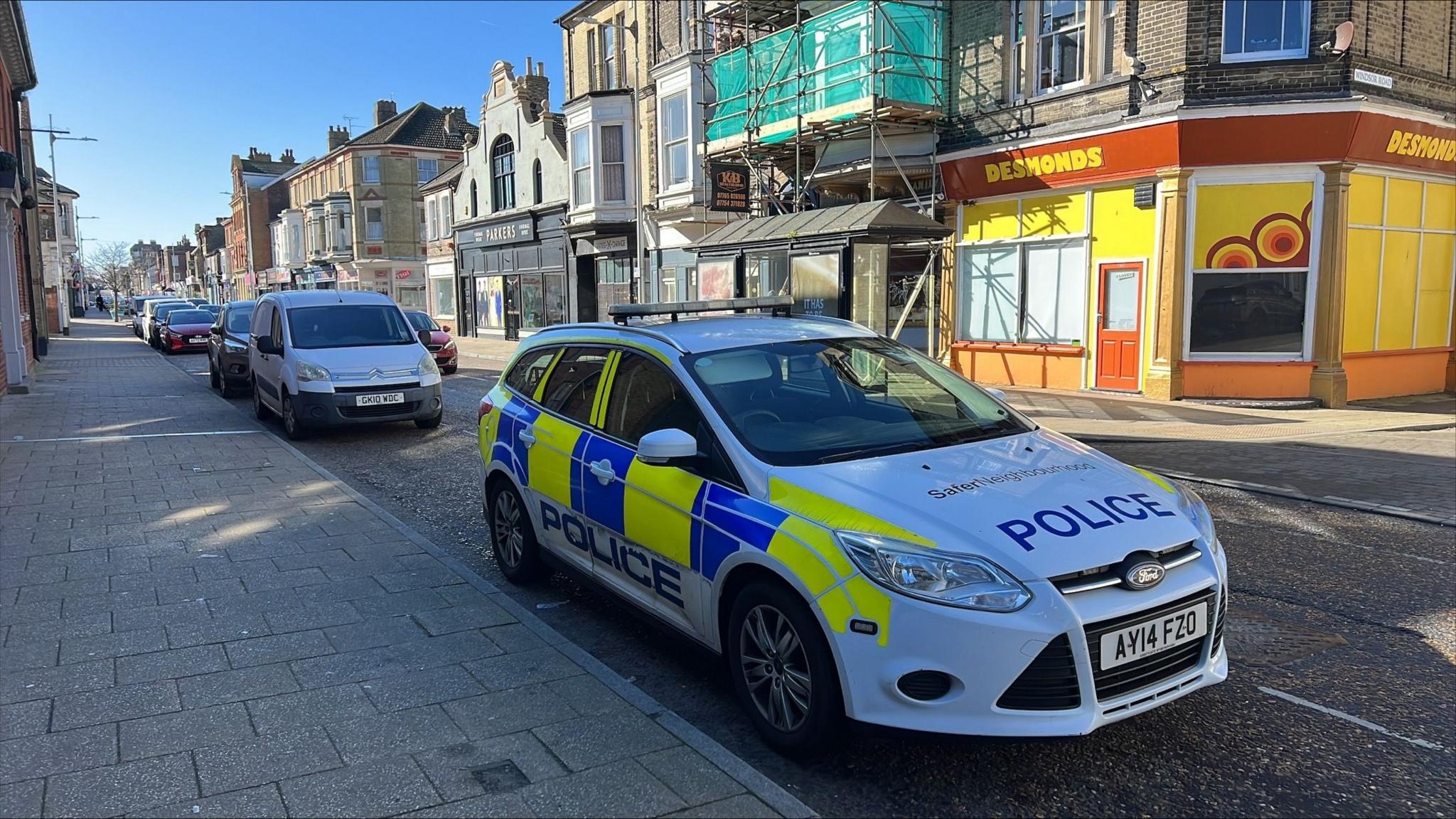 A police car is parked up on London Road South in Lowestoft. Other vehicles are parked behind it. Buildings can be seen on the opposite side of the road some with scaffolding on them.