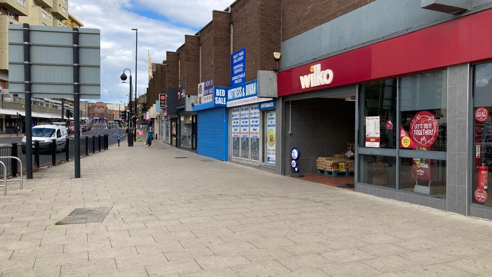 A shopping high street with a row of shops. The street is broadly empty, but for street furniture and one pedestrian in the far distance.