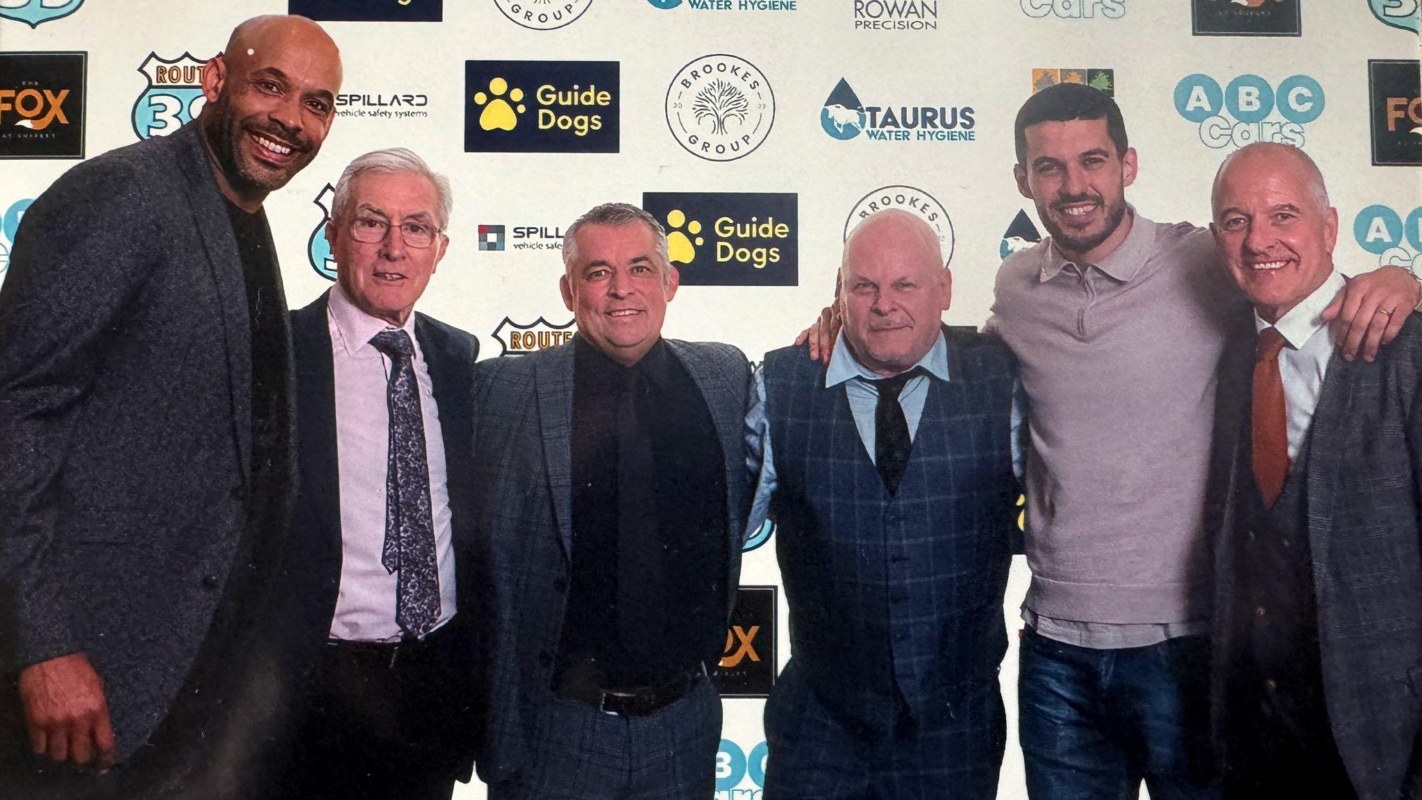 Six men, arms around shoulders, in front of a sponsors' board