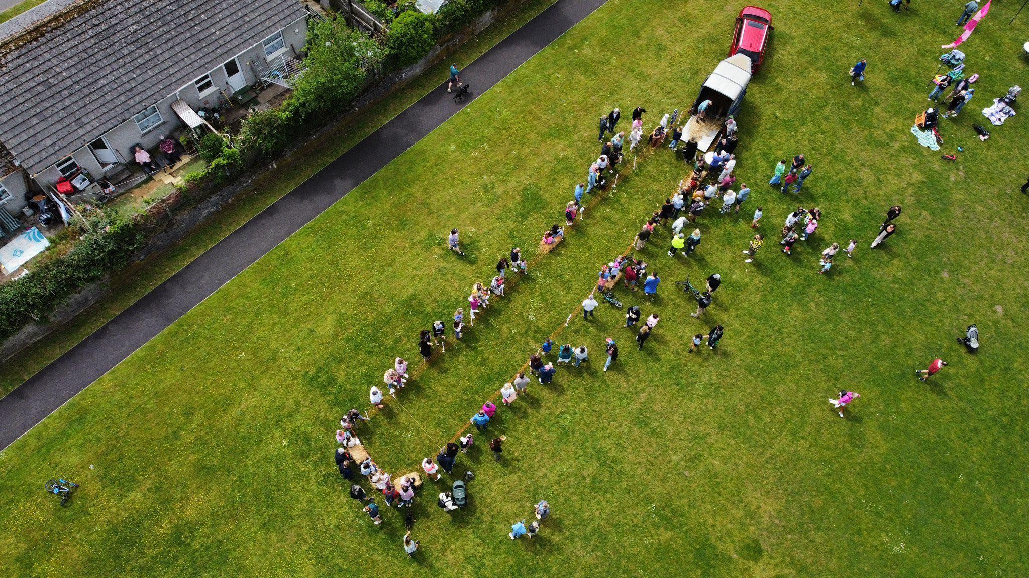 Aerial view of a large group of people standing in a winding line on a grassy field. A red vehicle with an open trailer is parked in the top right corner, with several people nearby. In the background, a row of houses with backyards lines the edge of the field, and a paved path runs parallel to the houses with a few individuals walking along it.
