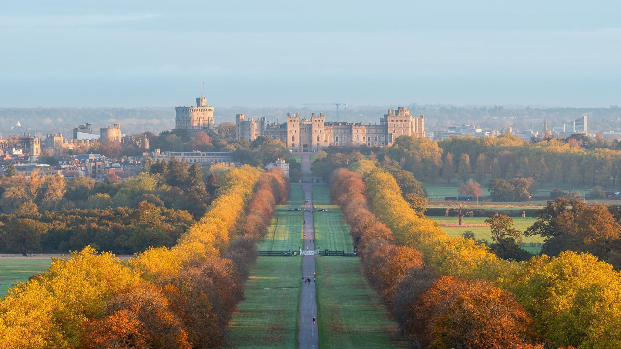 Windsor Castle dominates the skyline of this picture, the stone glowing in the sun. The long walk runs up to the castle, with people walking on the path. The trees each side are beautiful shades of orange, brown and yellow. the sky overhead is blue with light cloud. Behind the castle you can see buildings stretching away on the hillside.