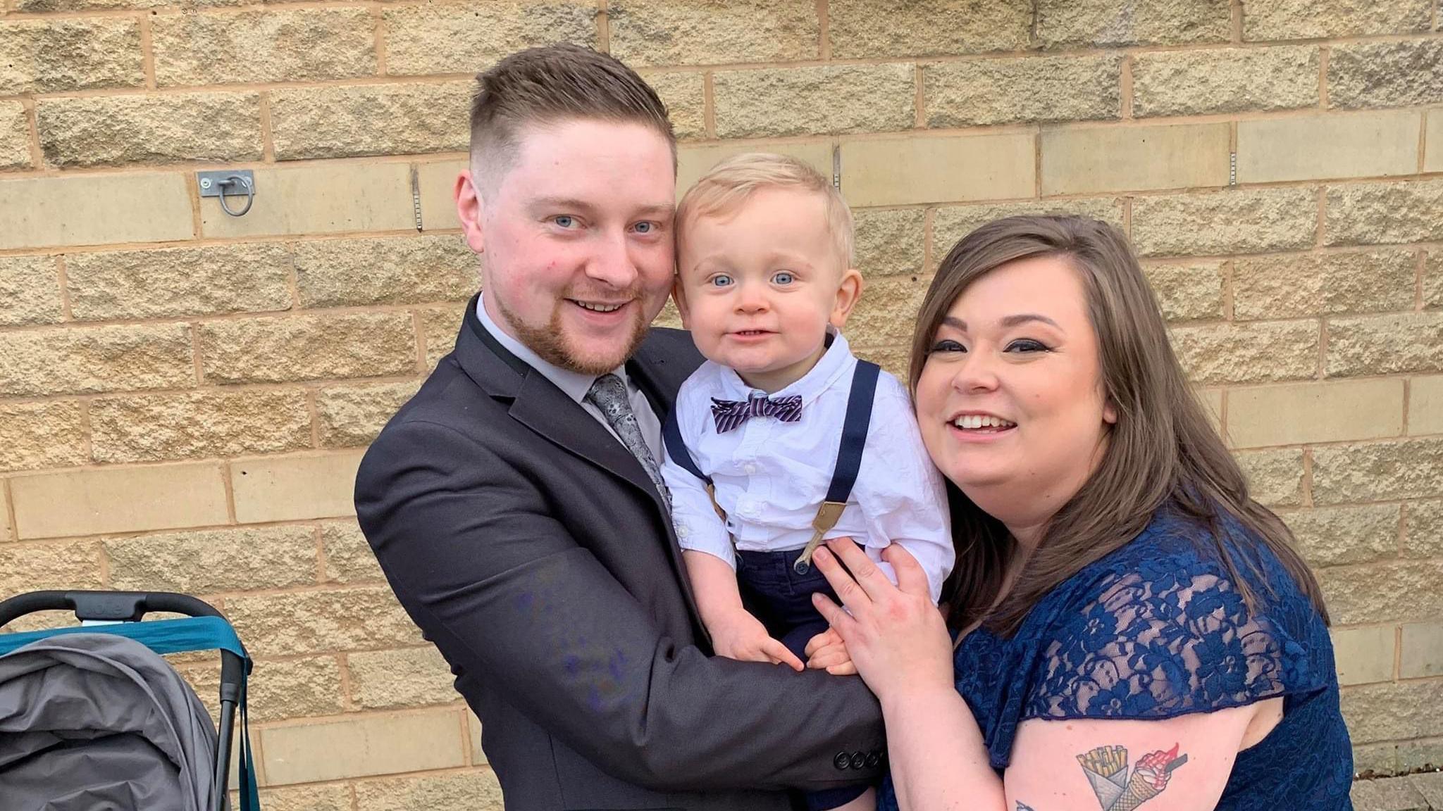 A man in a suit and a woman in a blue lacy dress stand holding a young boy in between them and smiling at the camera. They are standing in front of a yellow brick wall