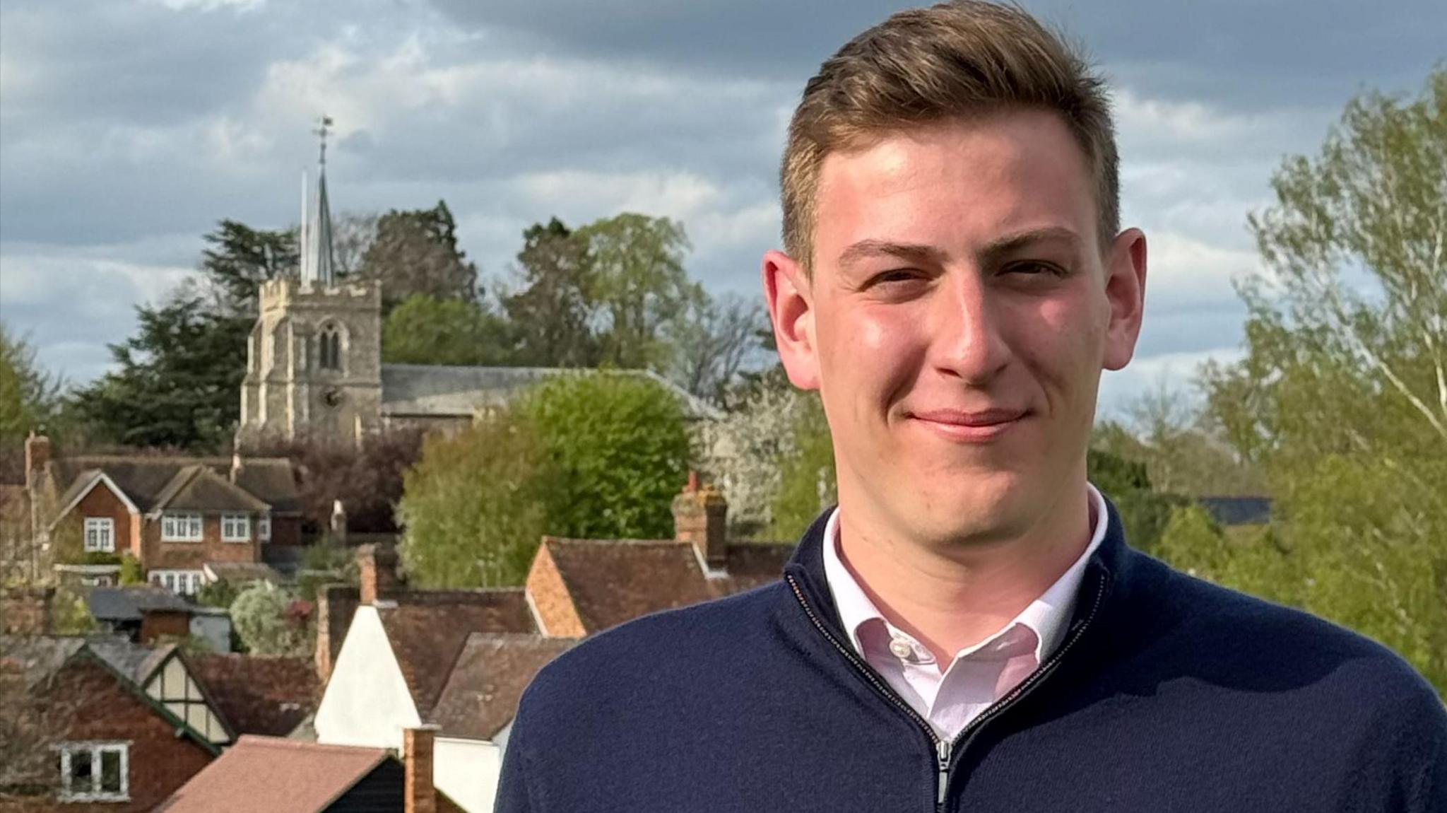 A man wearing a pink shirt and a blue quarter-zip top looking into the camera. He is clean shaven and has light brown hair. In the background there is a church, trees and houses.
