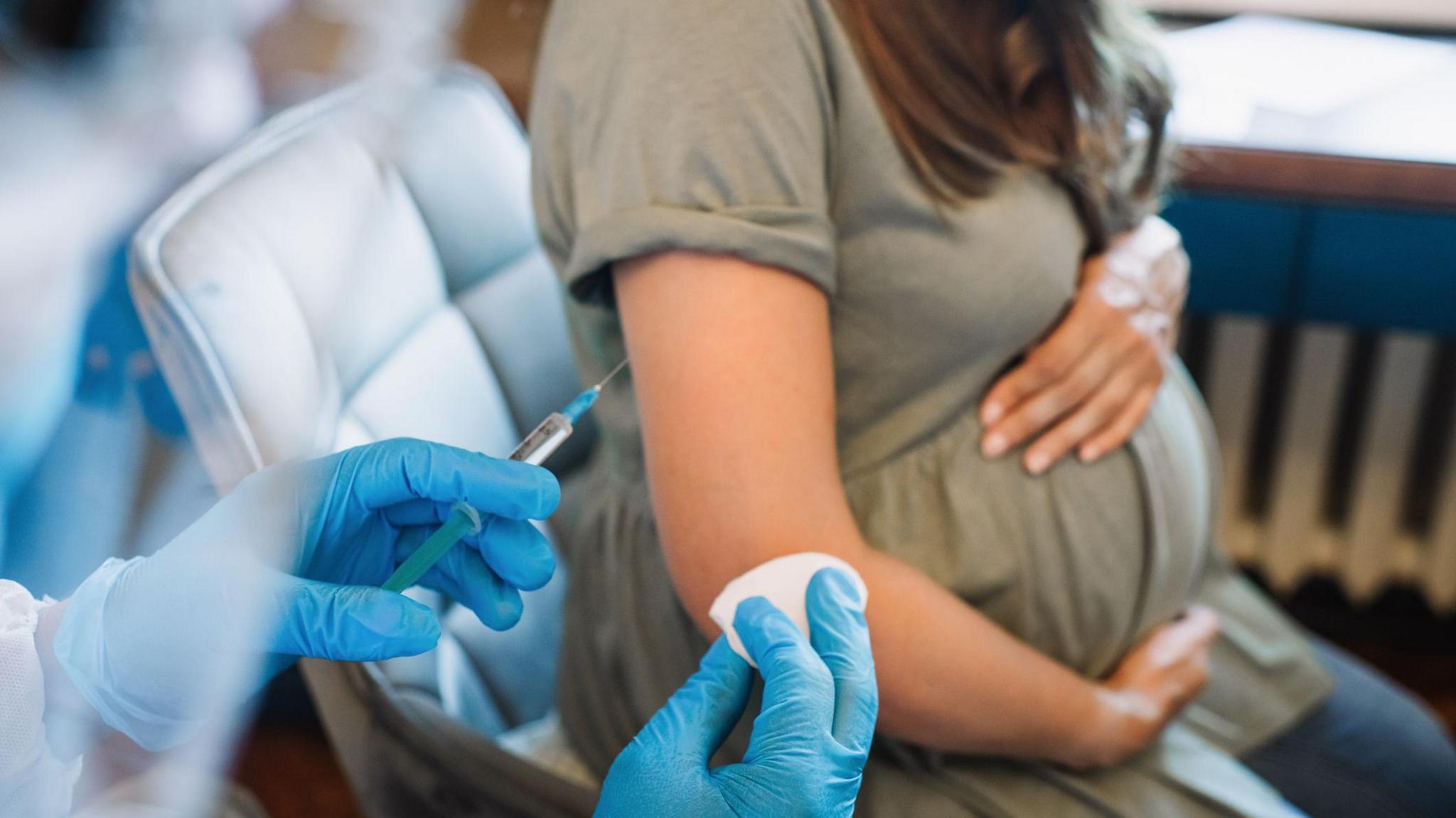 A photo of a medical professional wearing blue gloves holding a needle and cotton pad in front of a pregnant woman holding her baby bump