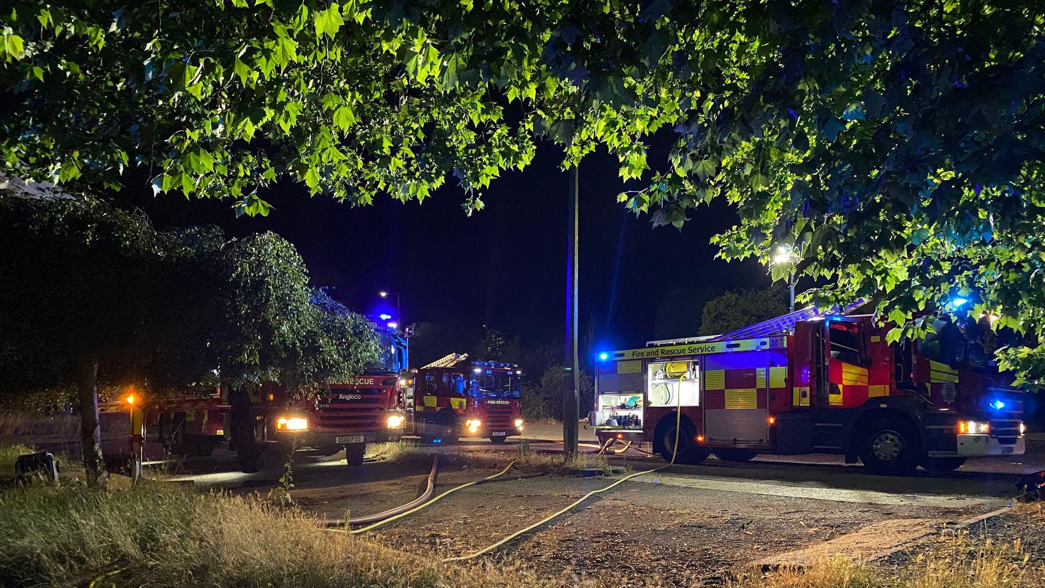 Three fire engines pictured in the dark with their headlights on. There are hoses running to them over muddy ground. There are trees in the foreground.